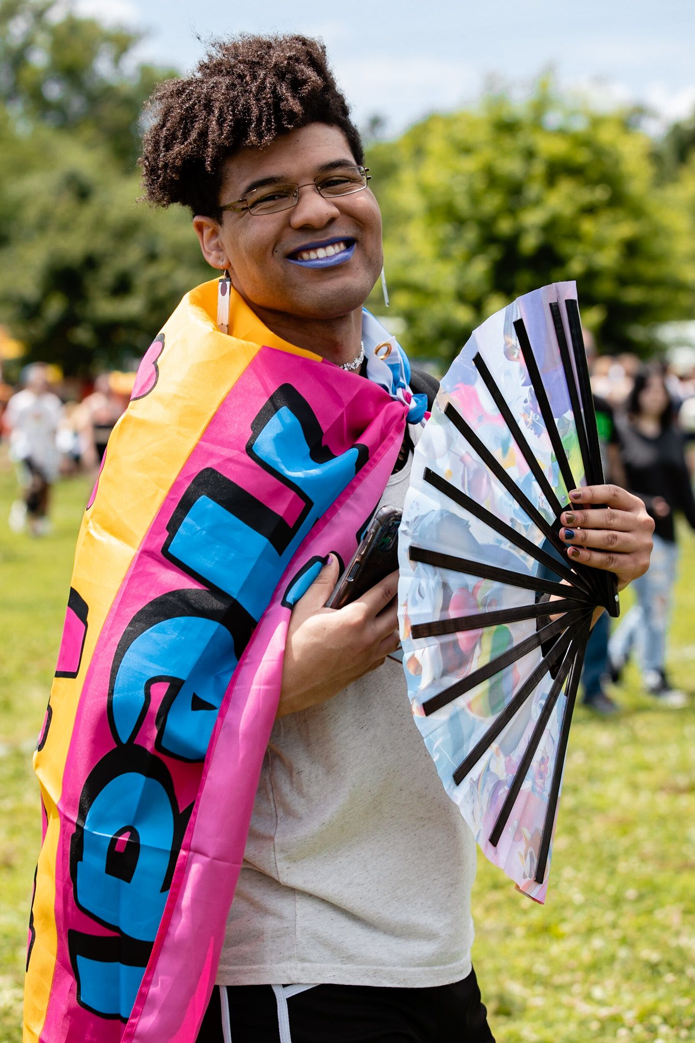 Person holding colorful pride fan while wearing vibrant pink and blue attire at outdoor event.