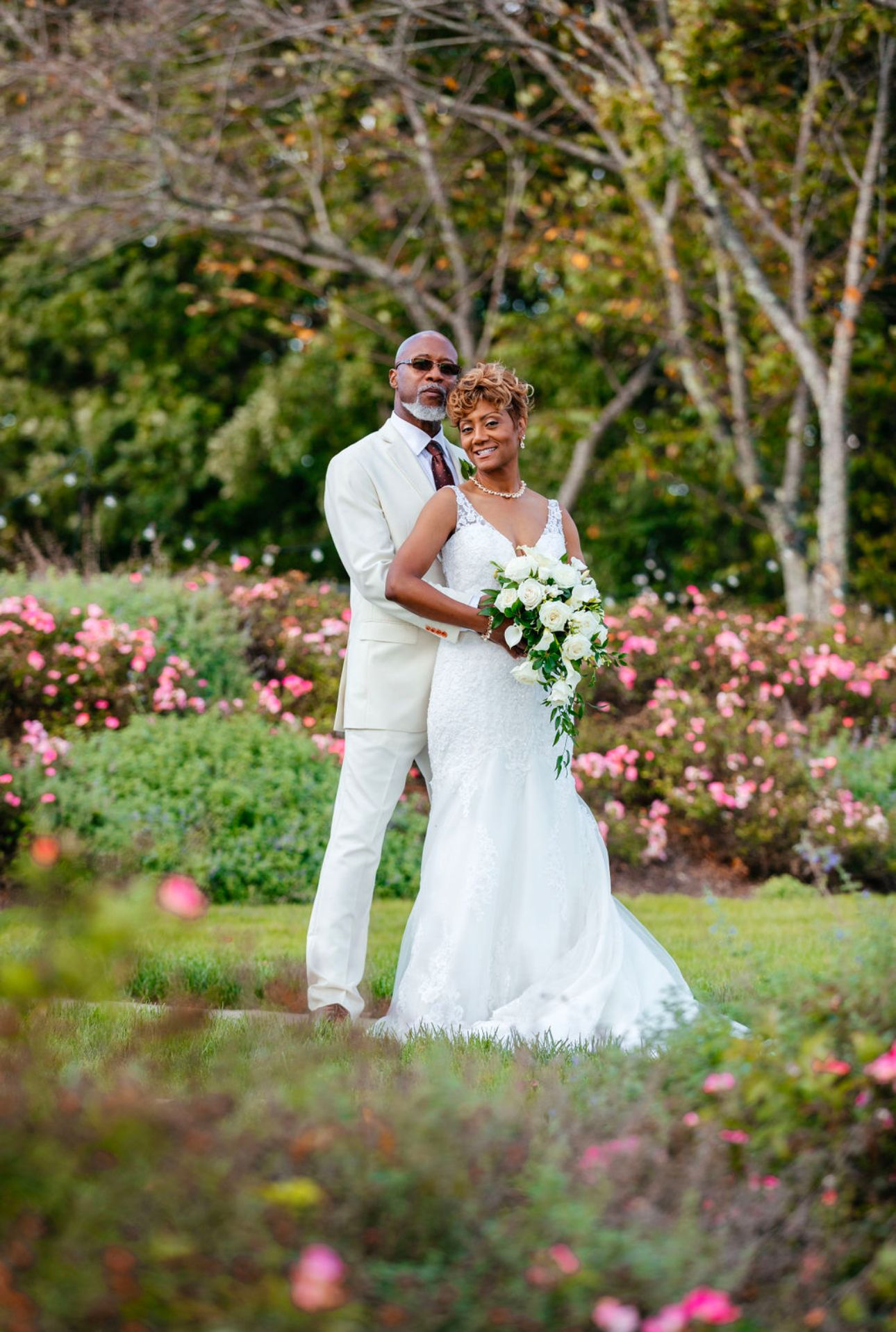 A couple in white wedding attire poses in a garden filled with pink roses and greenery.
