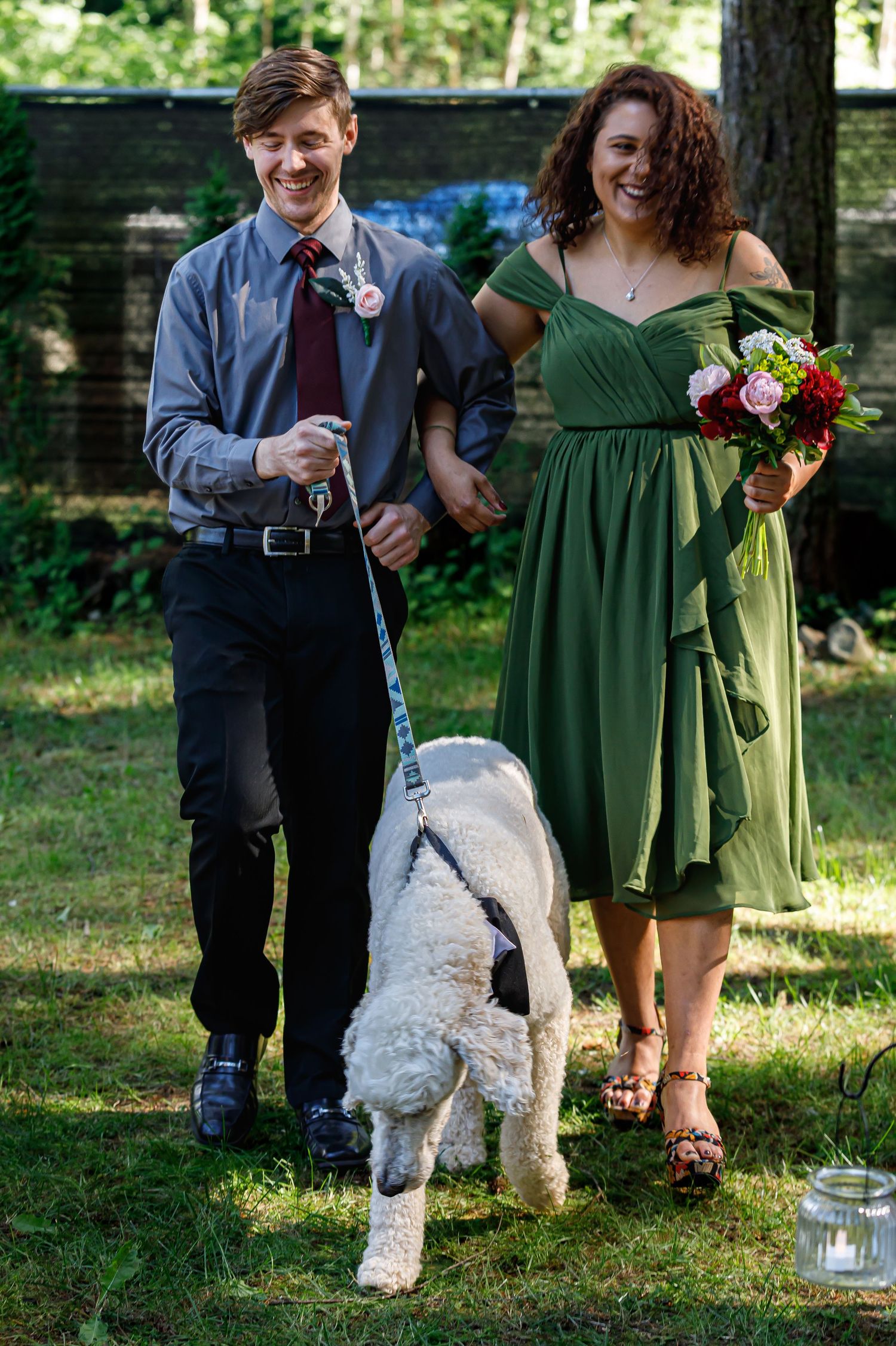 Wedding party members walk with a white dog on a leash while holding floral bouquets.