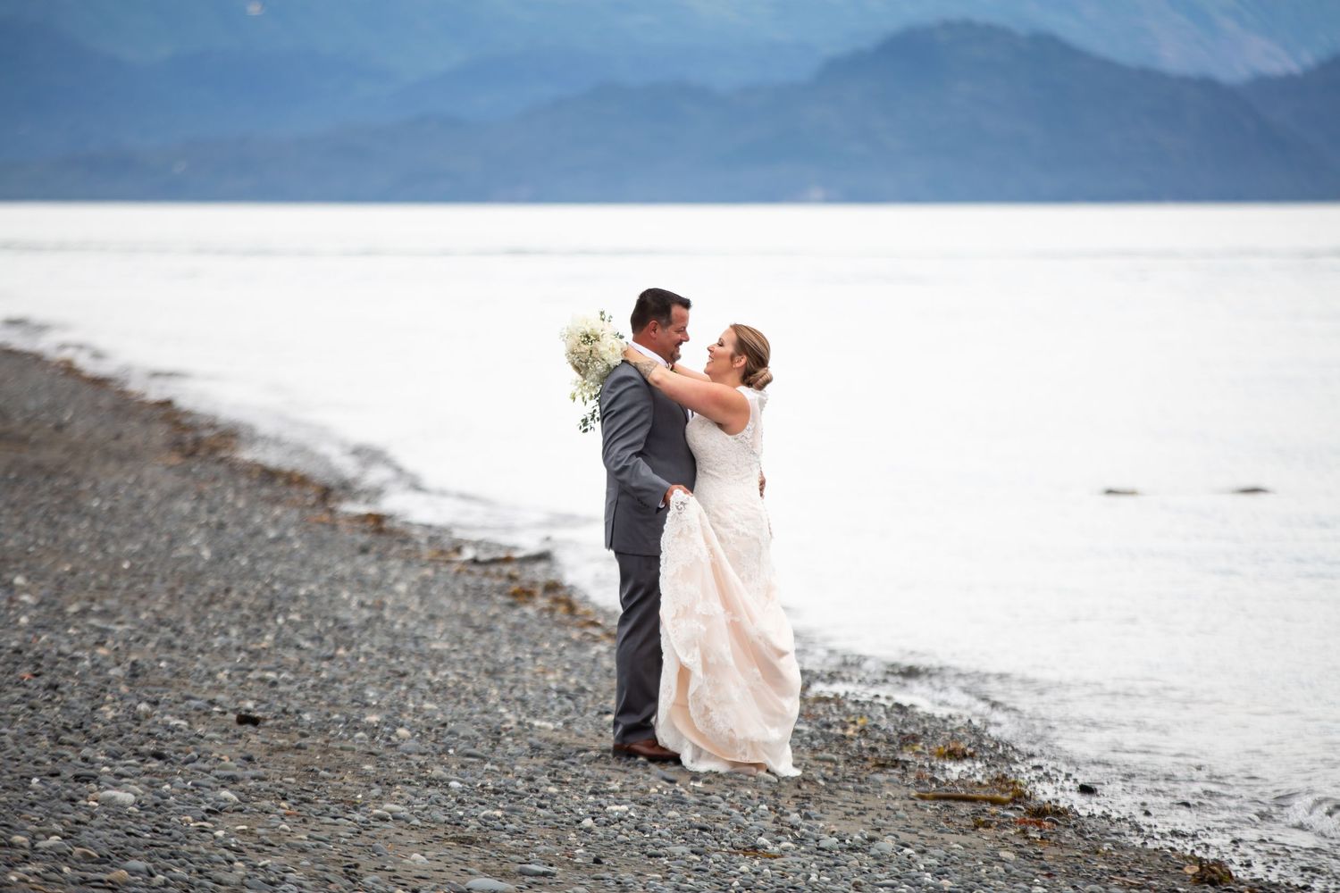 A wedding couple embraces on a rocky beach with mountains and water in the background.