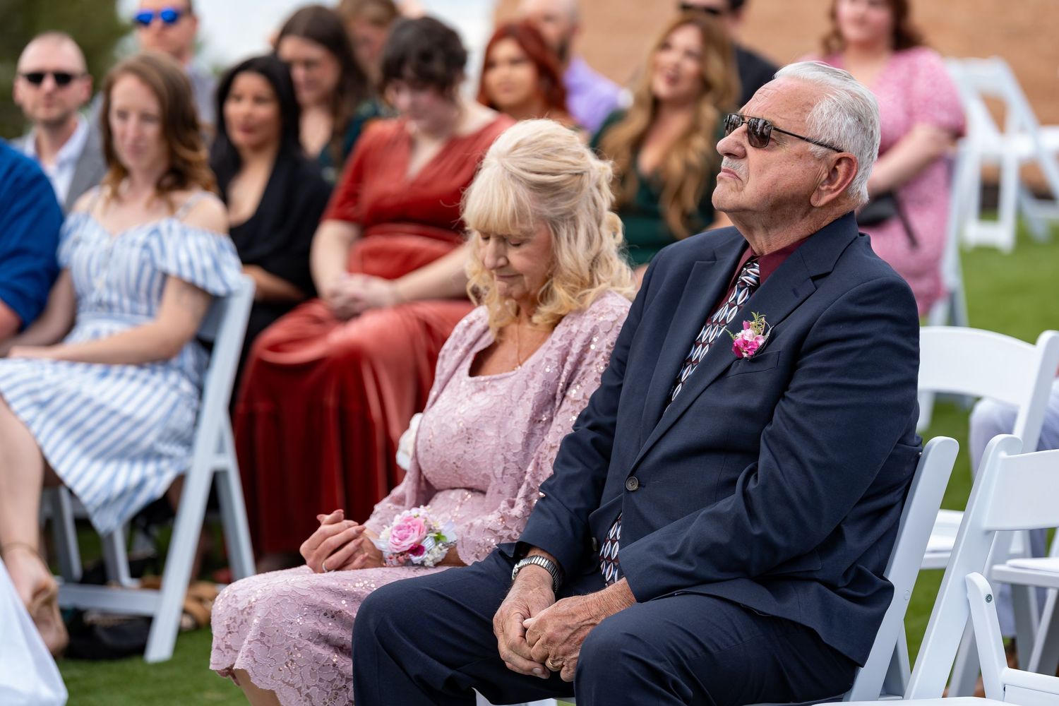 Wedding guests seated in white chairs at an outdoor ceremony watching the proceedings intently.