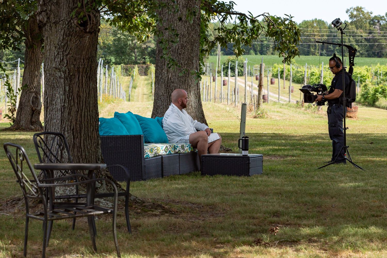 Outdoor seating area under large trees with a groom relaxing on patio furniture being interviewed by a videographer on his wedding day.