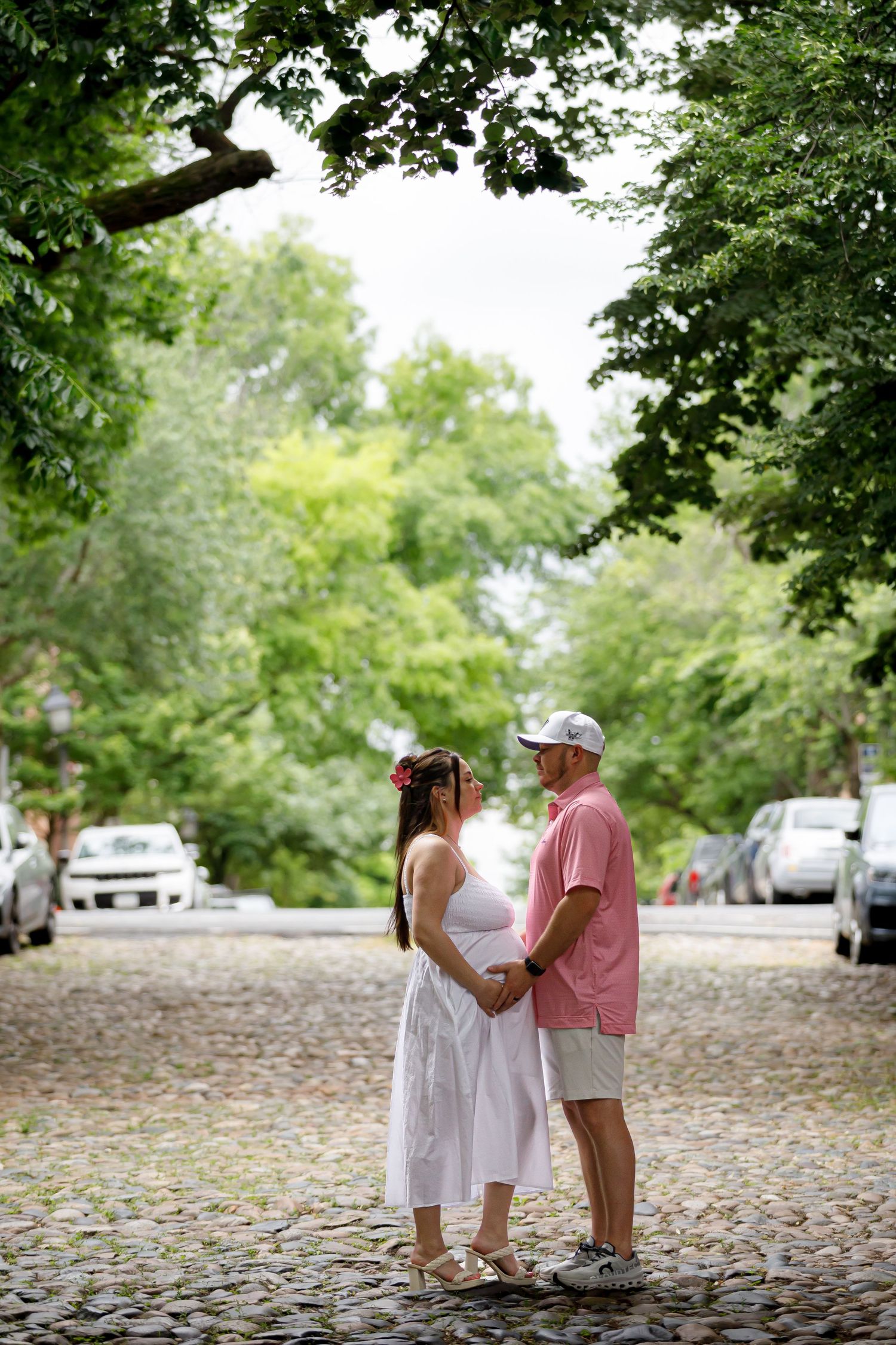 A couple poses for photos on a charming cobblestone street surrounded by lush green trees.