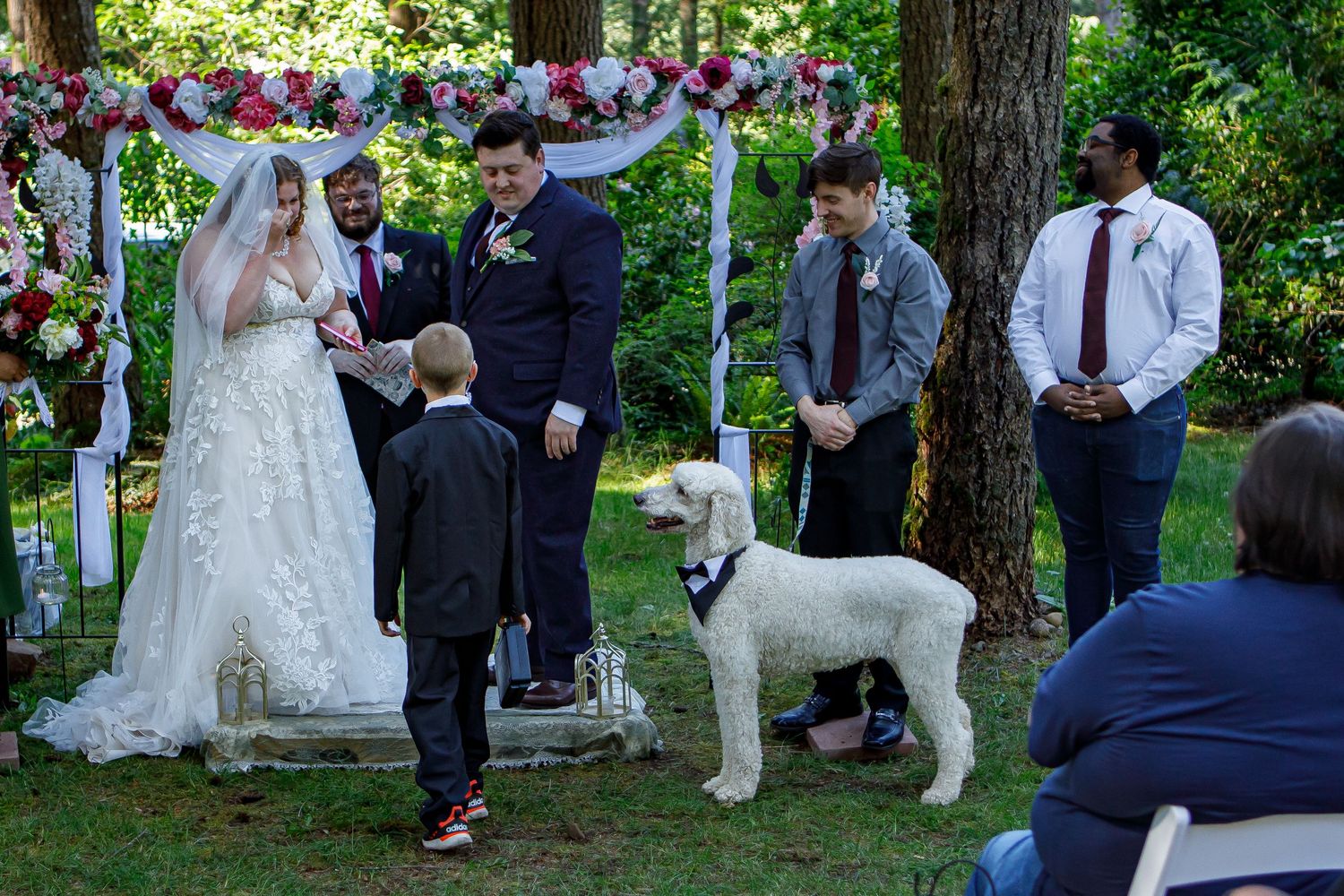 Outdoor wedding ceremony with guests seated on white chairs among trees.