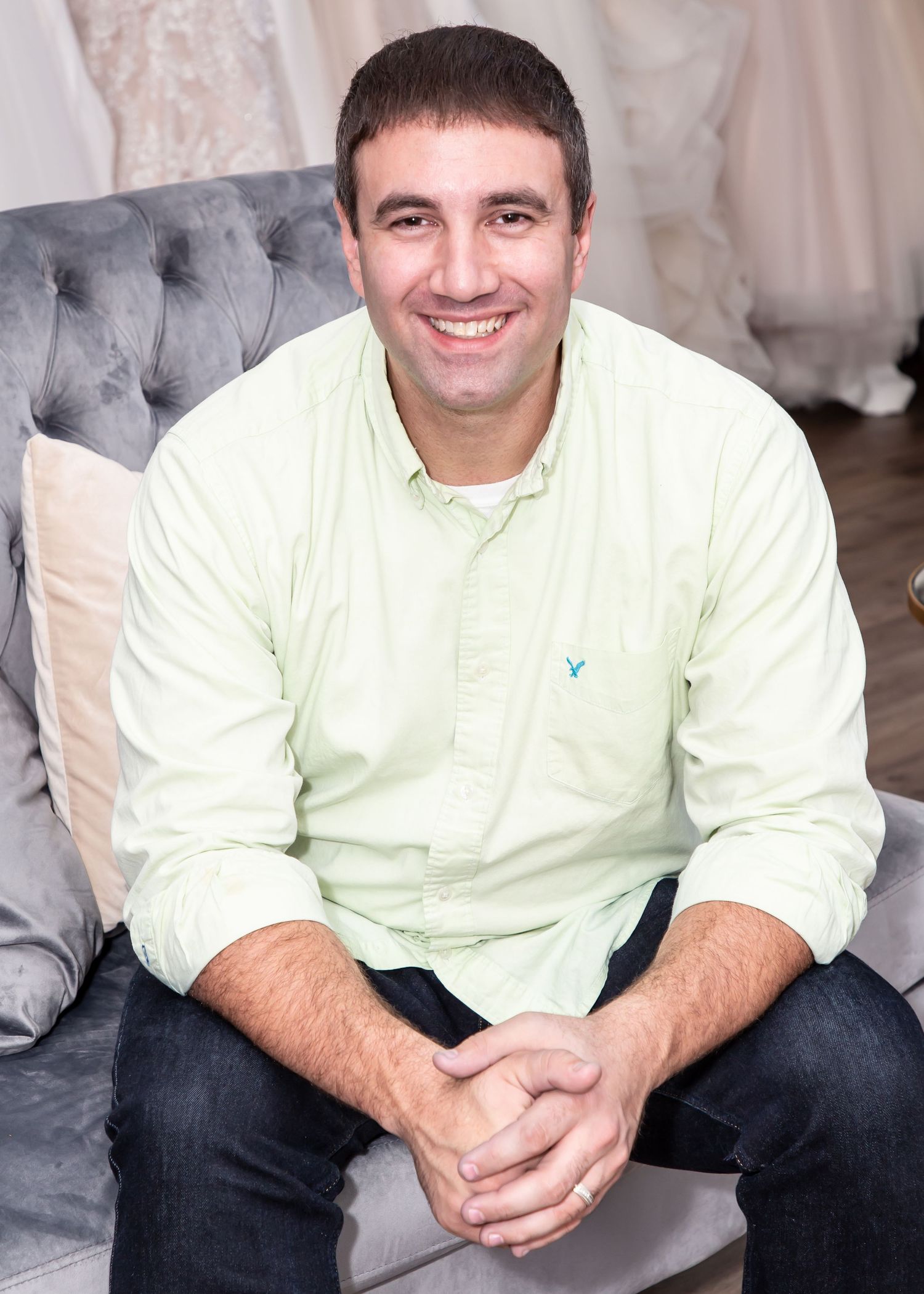 Man in light yellow shirt sitting on gray tufted couch in bridal shop.