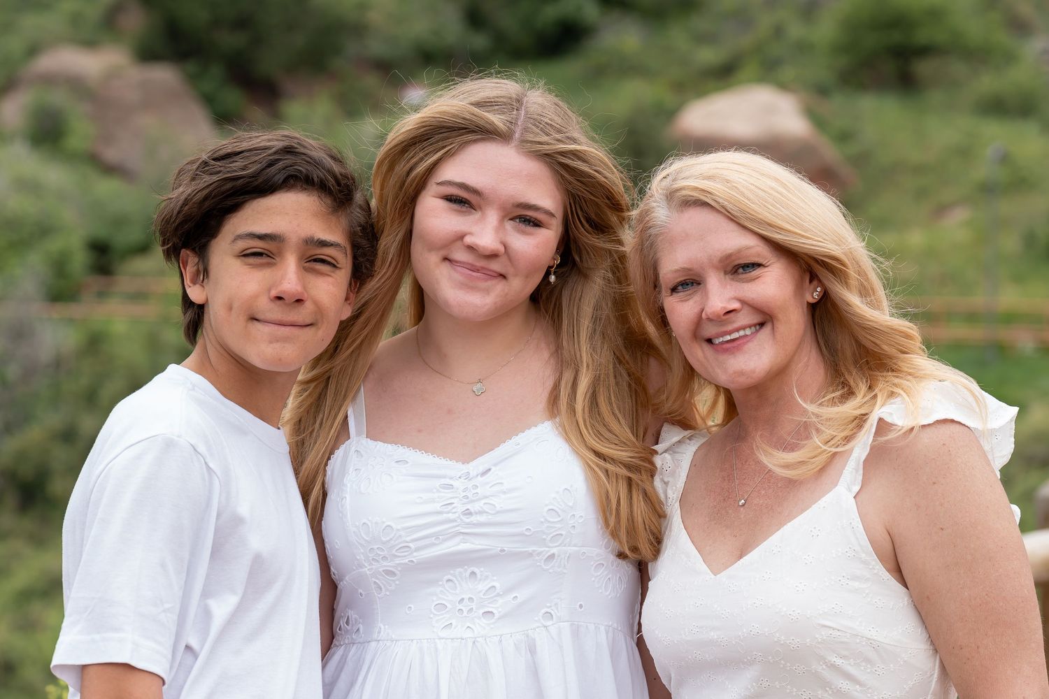 Three people wearing white outfits pose together outdoors against a mountain backdrop.