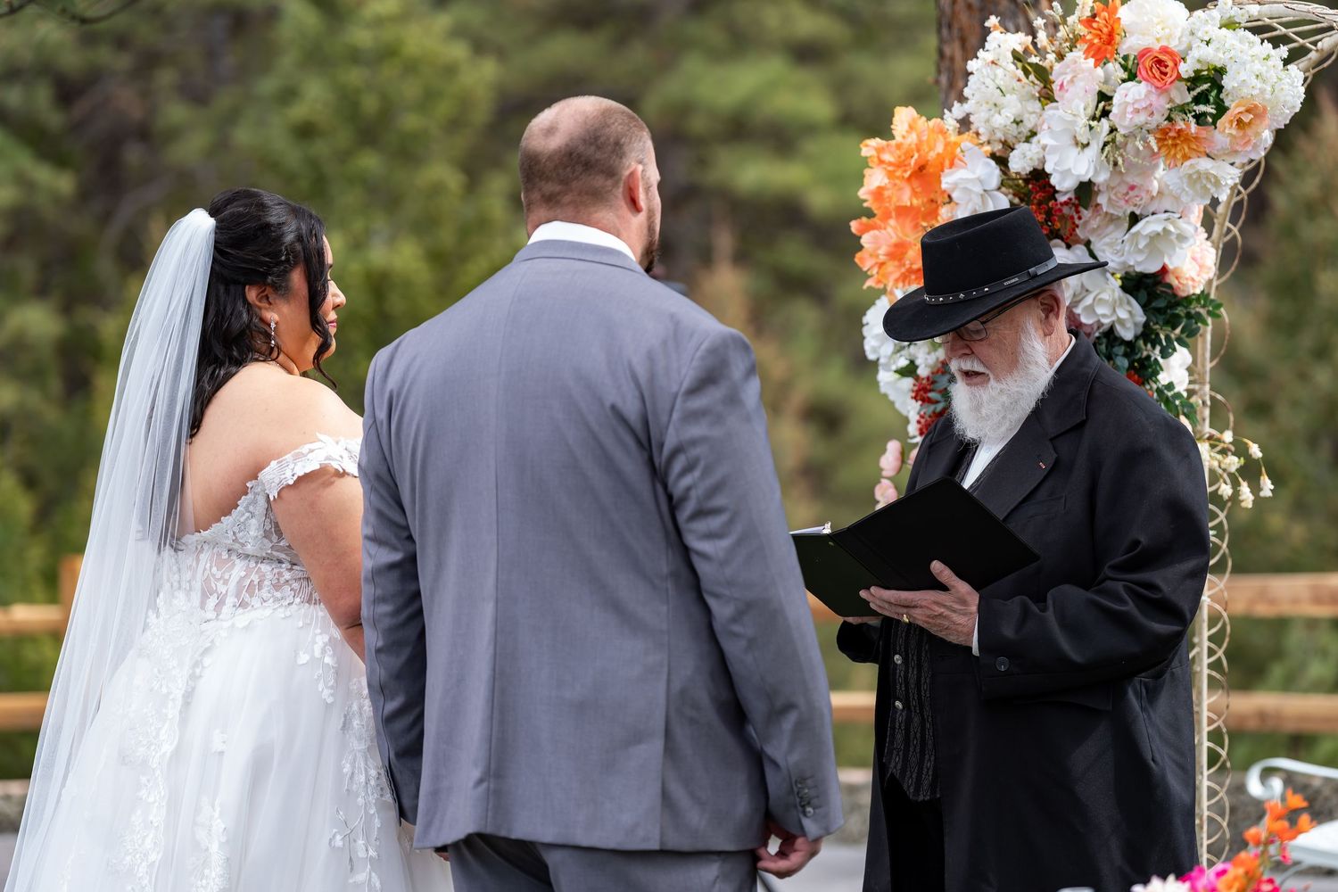 Wedding ceremony gathered around floral arch with orange and white flowers outdoors.