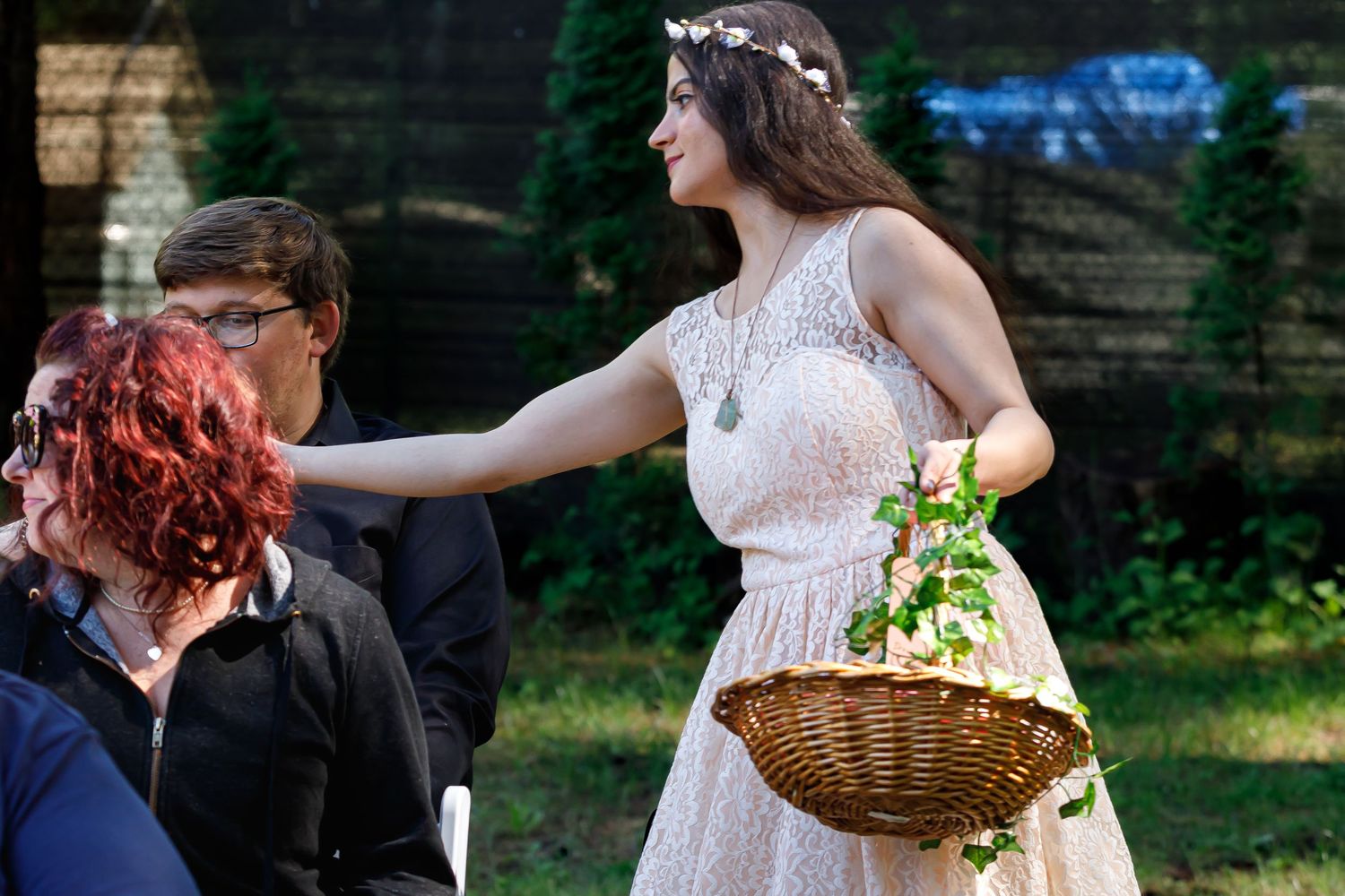 A flower girl in a white lace dress scatters petals and hands out cold drinks from a woven basket during an outdoor wedding ceremony.