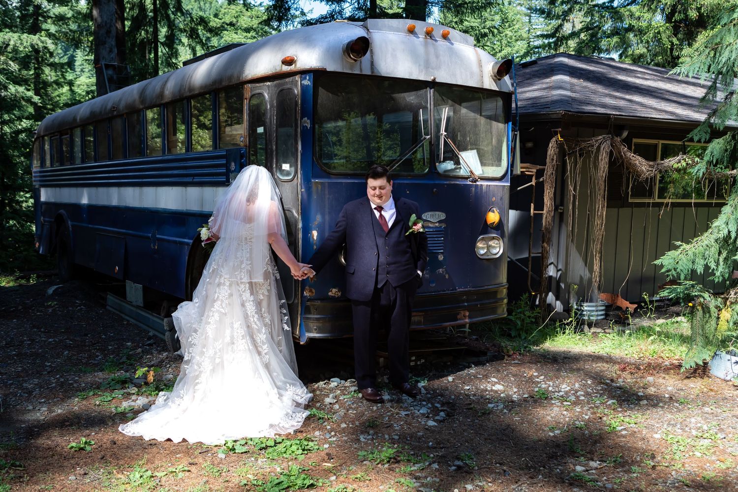 A couple poses for wedding photos next to a classic blue bus surrounded by greenery.