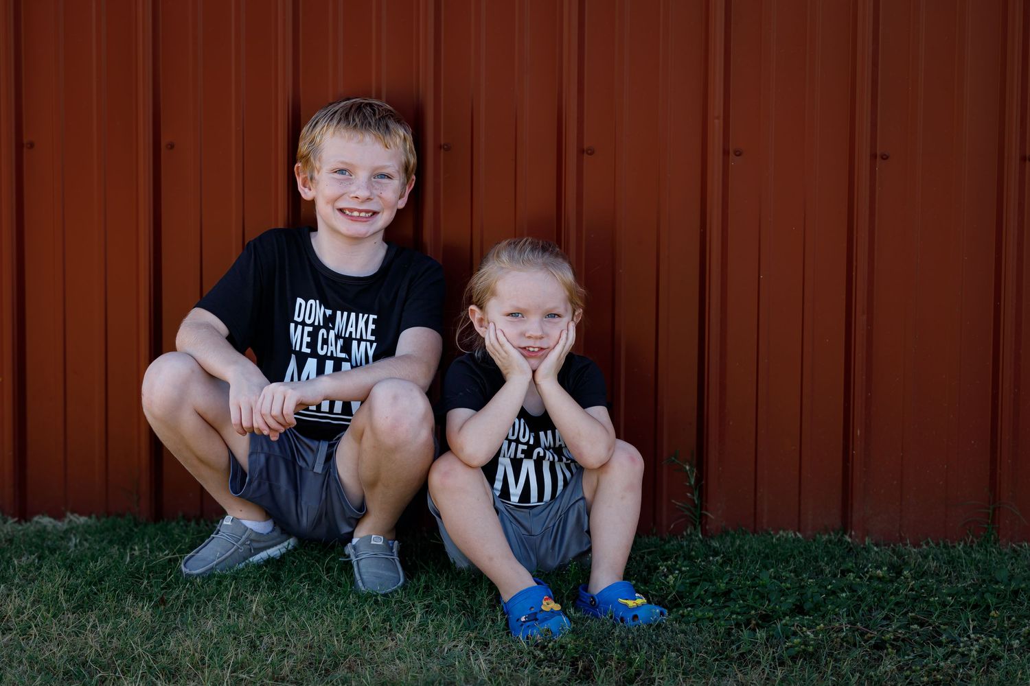 Children sitting and smiling against red metal wall wearing matching outfits.