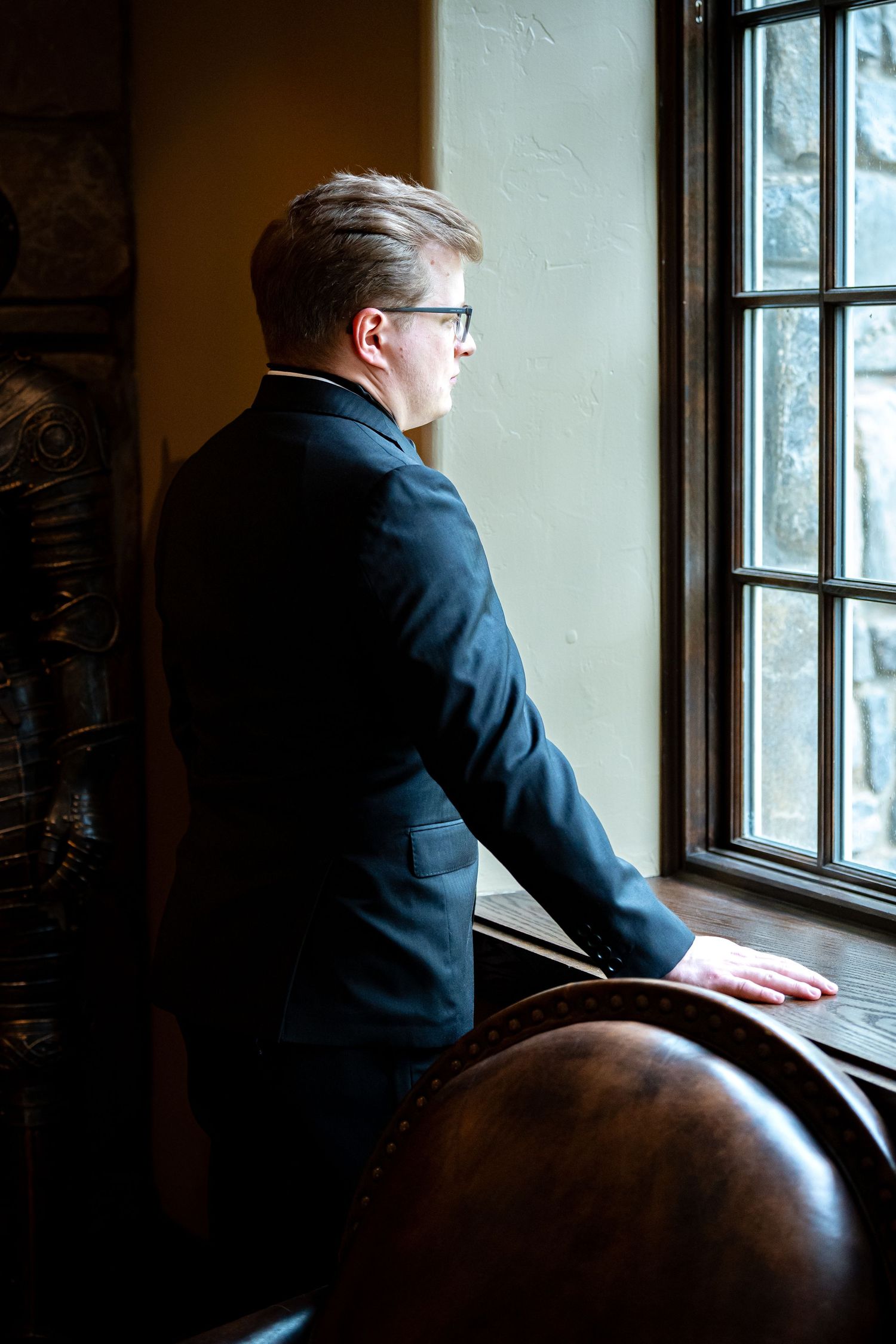 Groom in navy suit gazes thoughtfully out of window in moody lighting.