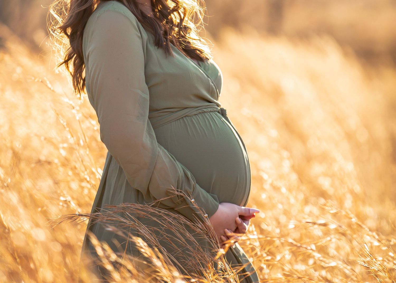 Silhouette of a pregnant woman in flowing sage green dress standing in golden wheat field at golden hour sunset.