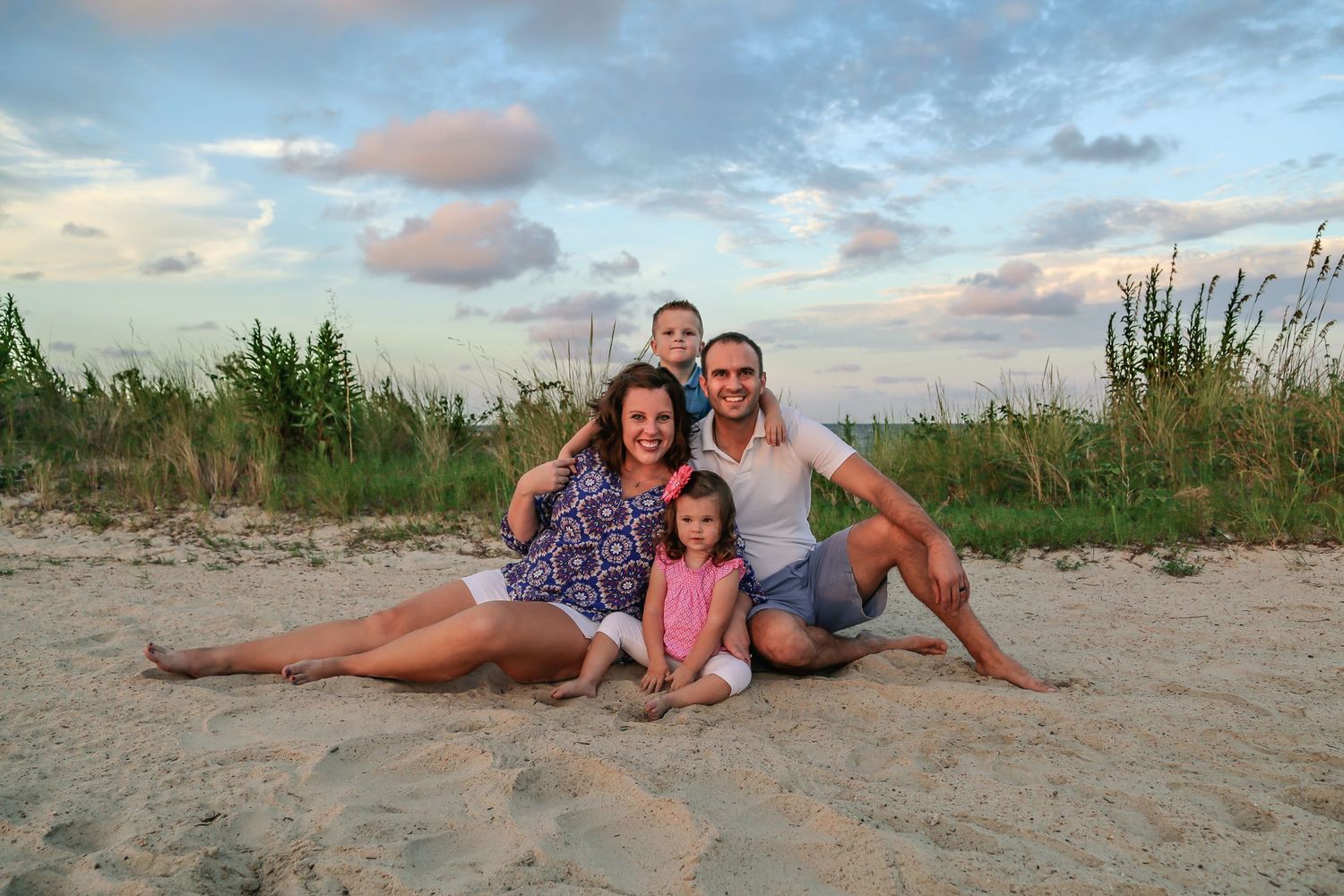Happy family sitting together on beach in Virginia Beach at sunset with coastal vegetation and beautiful sunset sky in background.