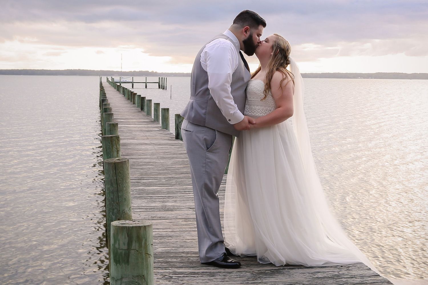 A romantic couple shares a tender moment on a wooden pier extending into calm waters at sunset.