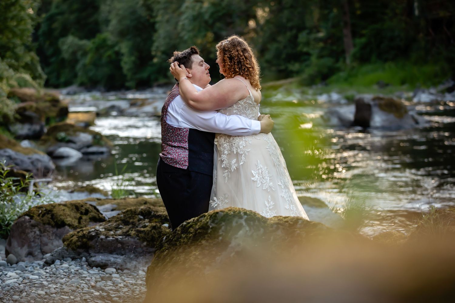 A romantic wedding couple embraces by a flowing river surrounded by lush forest.