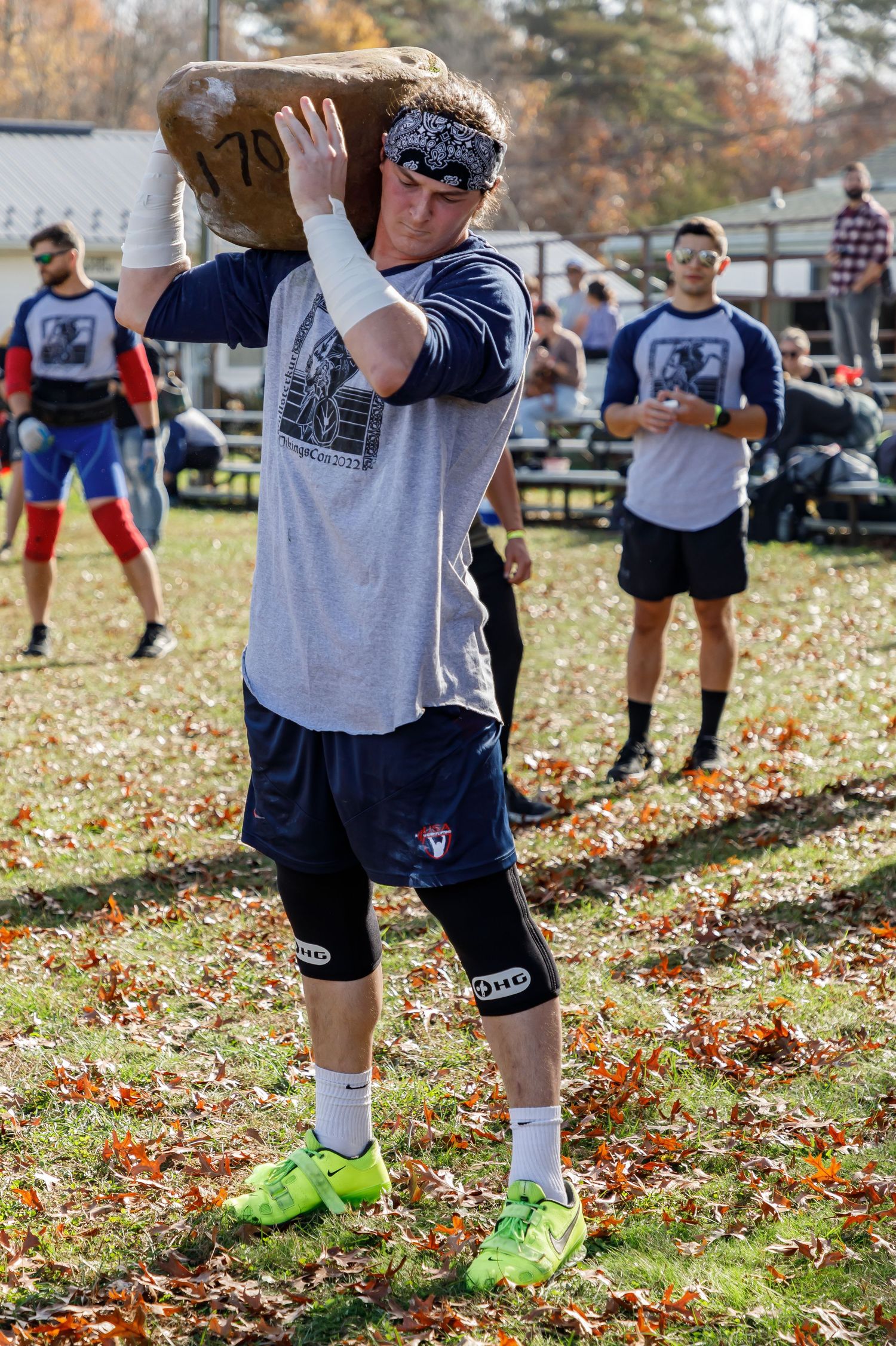 Athletes in matching t-shirts compete in outdoor log lifting event on fallen autumn leaves.