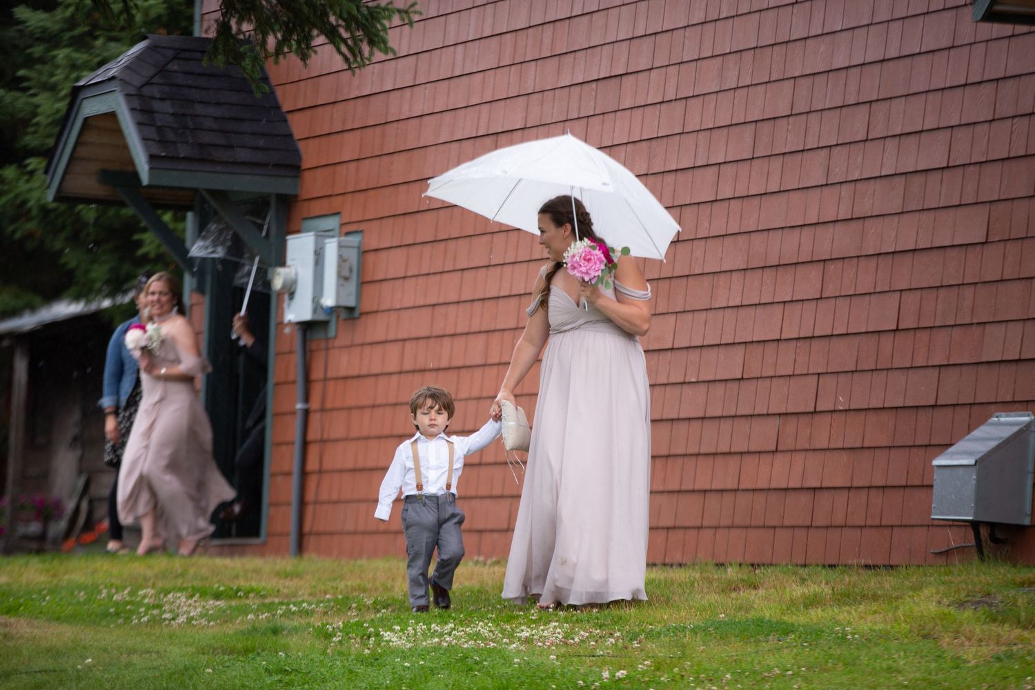 Wedding party members with umbrellas line up against a cedar-shingled building.