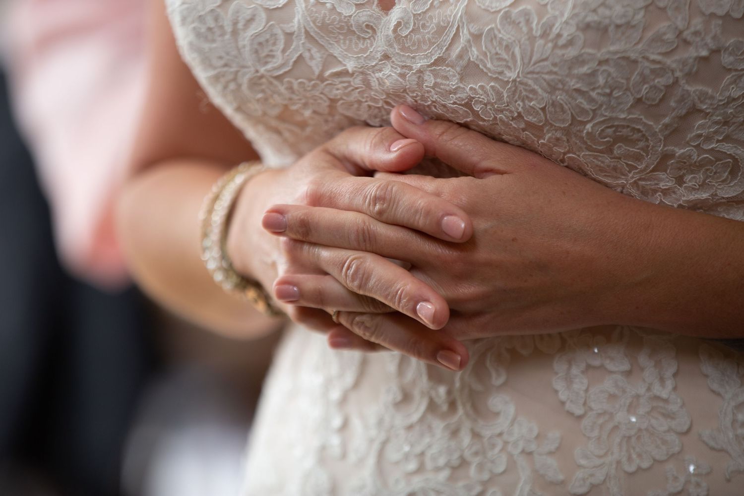 Close-up details of a white lace wedding dress with folded hands.