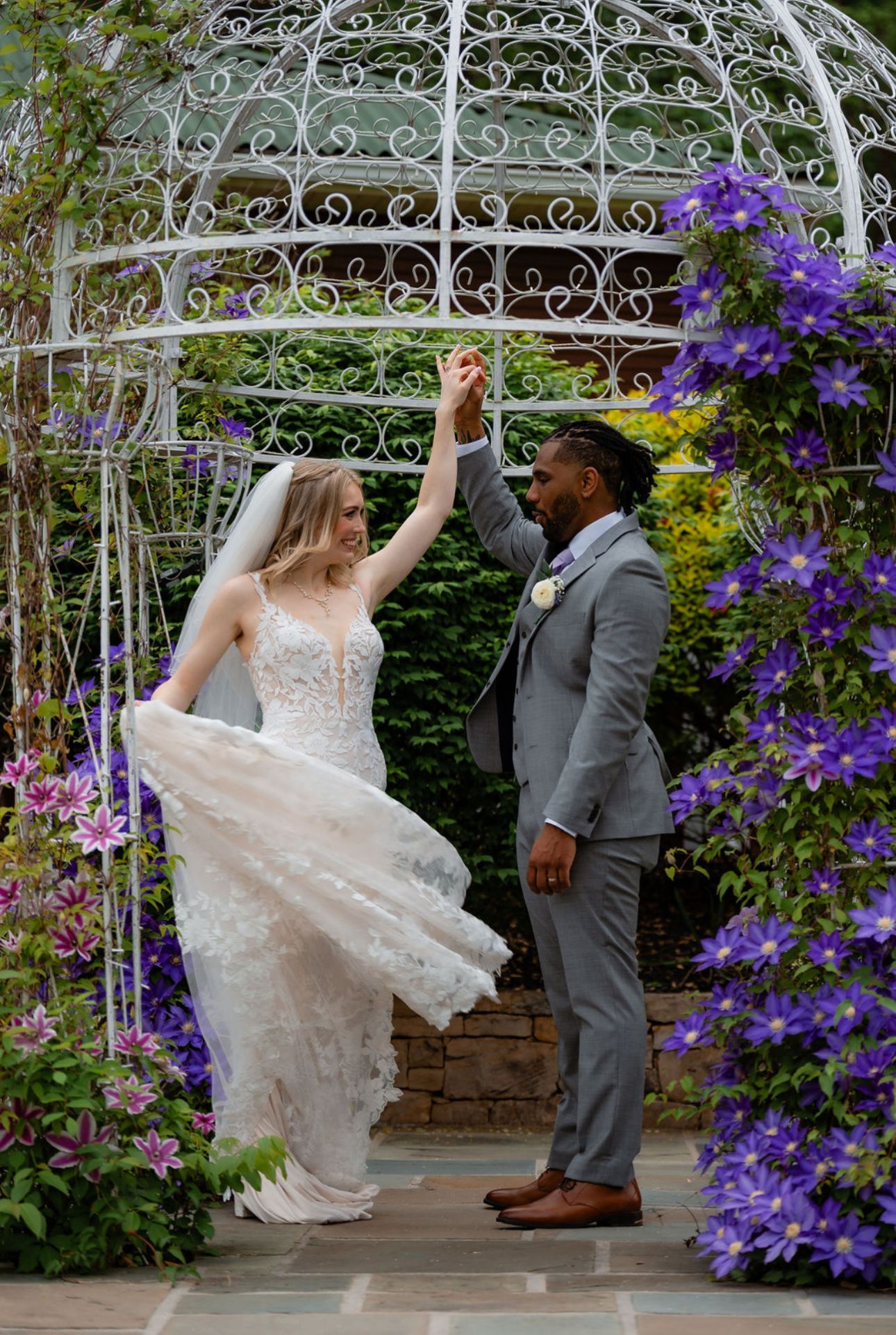 A couple dances under an ornate white gazebo decorated with vibrant purple clematis flowers in a garden setting.