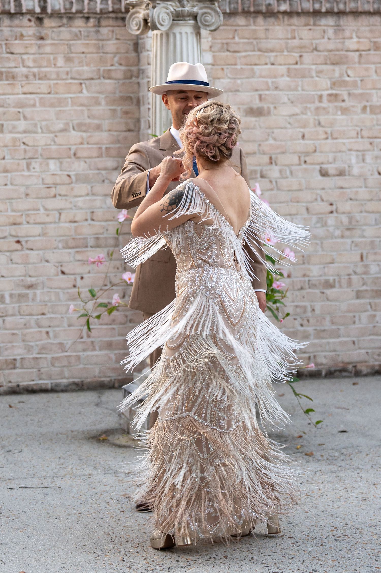 Dancing couple in vintage-inspired attire with flowing fringe dress against brick wall.