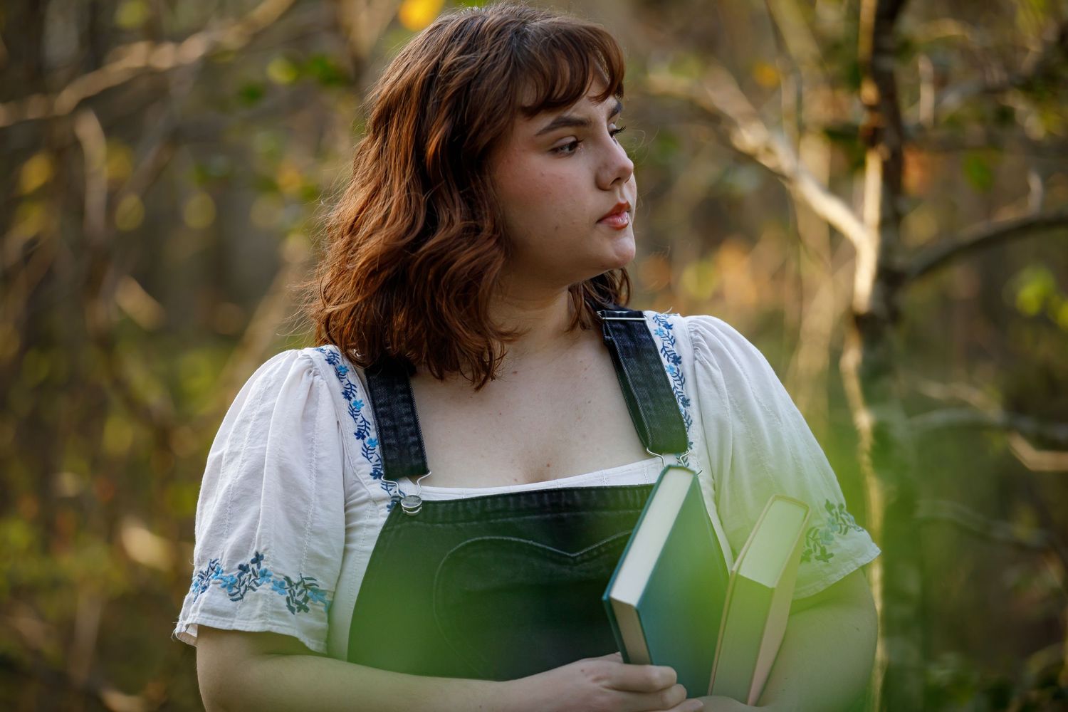 Person in green overalls sits thoughtfully among foliage during an outdoor photo session.