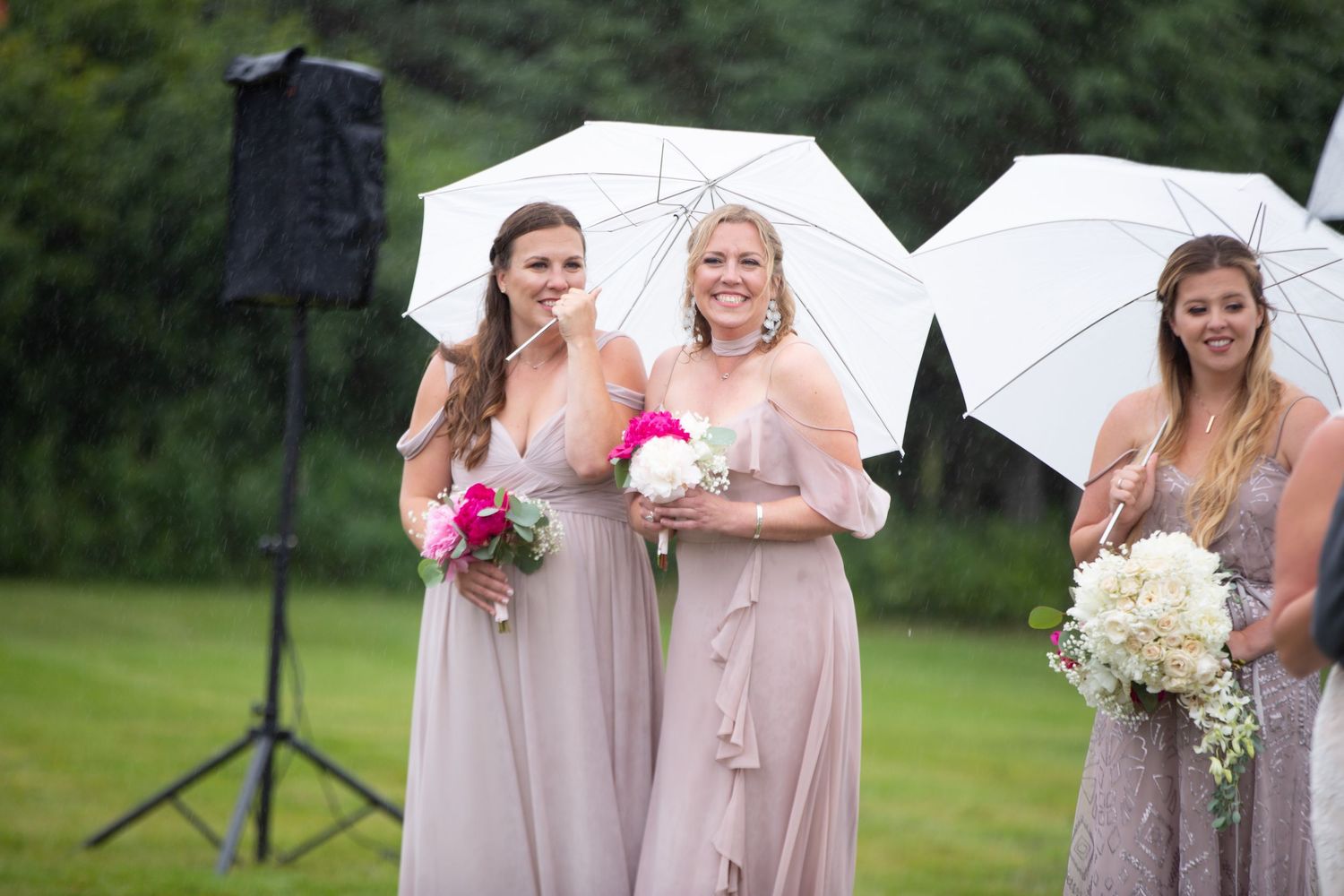 Wedding party members in blush pink dresses hold white umbrellas and bouquets on a grassy lawn.