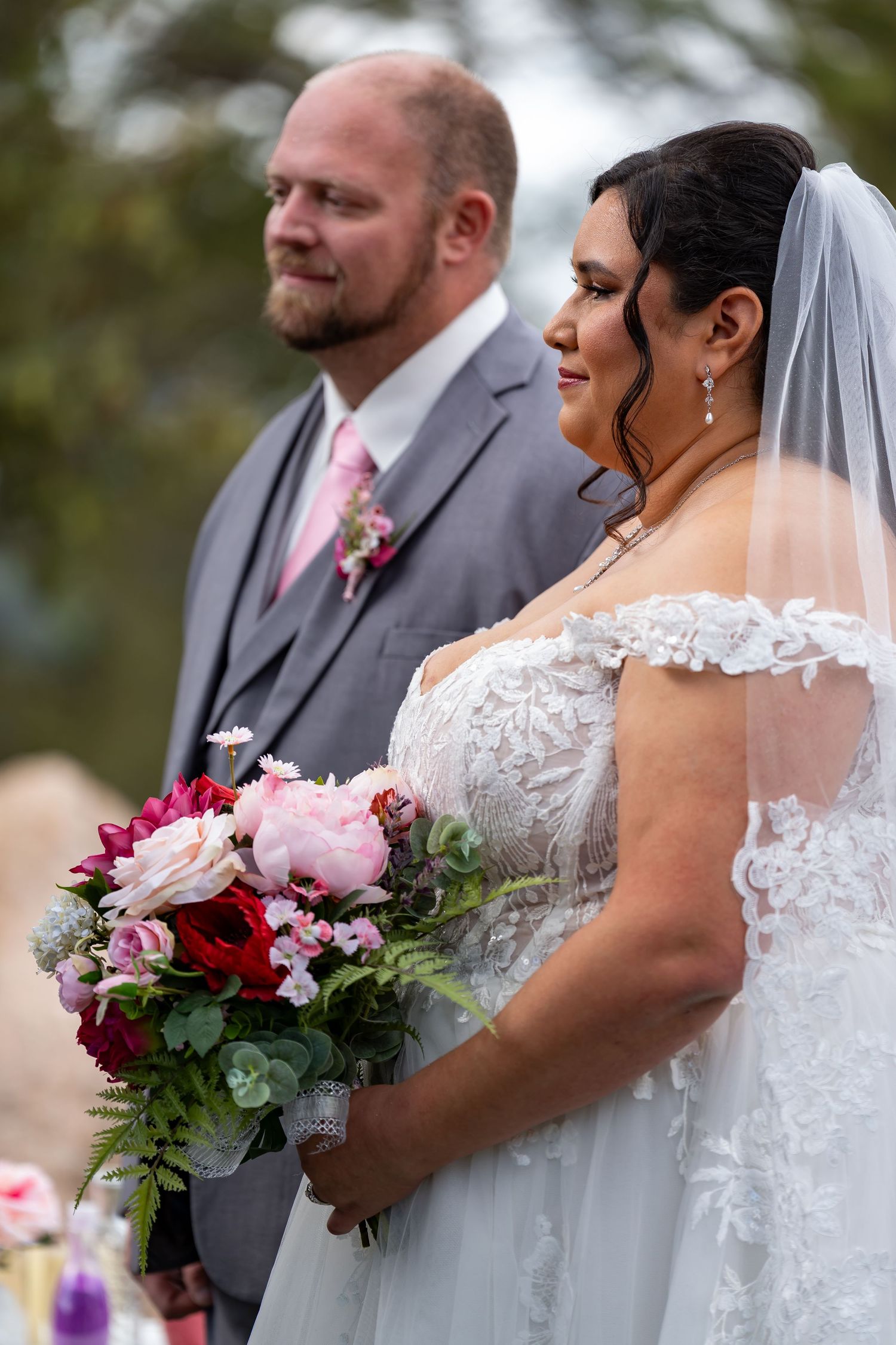 Bride in lace white dress holds colorful bouquet with pink and red flowers during wedding ceremony.