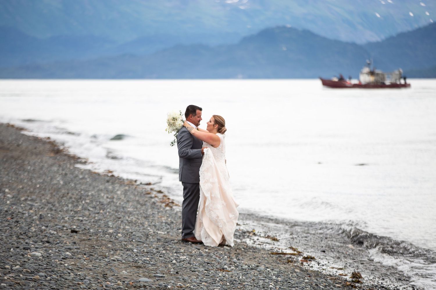 Bride and groom couple embracing on a snowy beach in Homer, Alaska with a fishing ship visible in the distance.