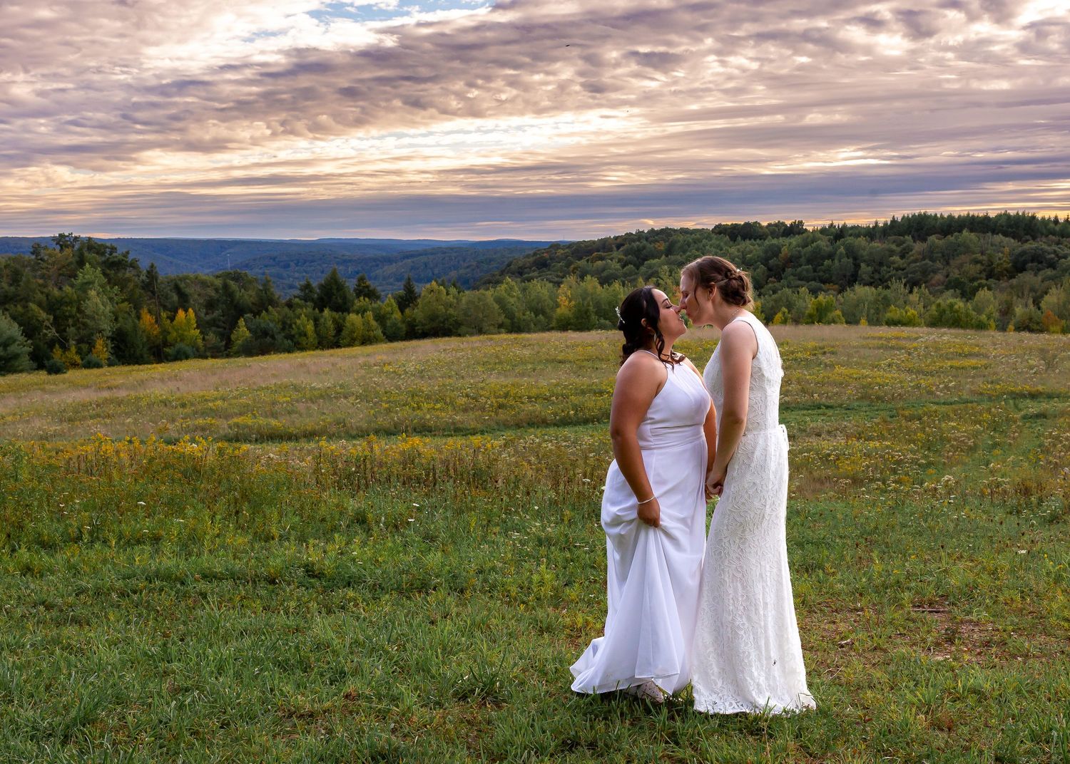 Two brides in white dresses stand together in a field at sunset with mountains in the background.