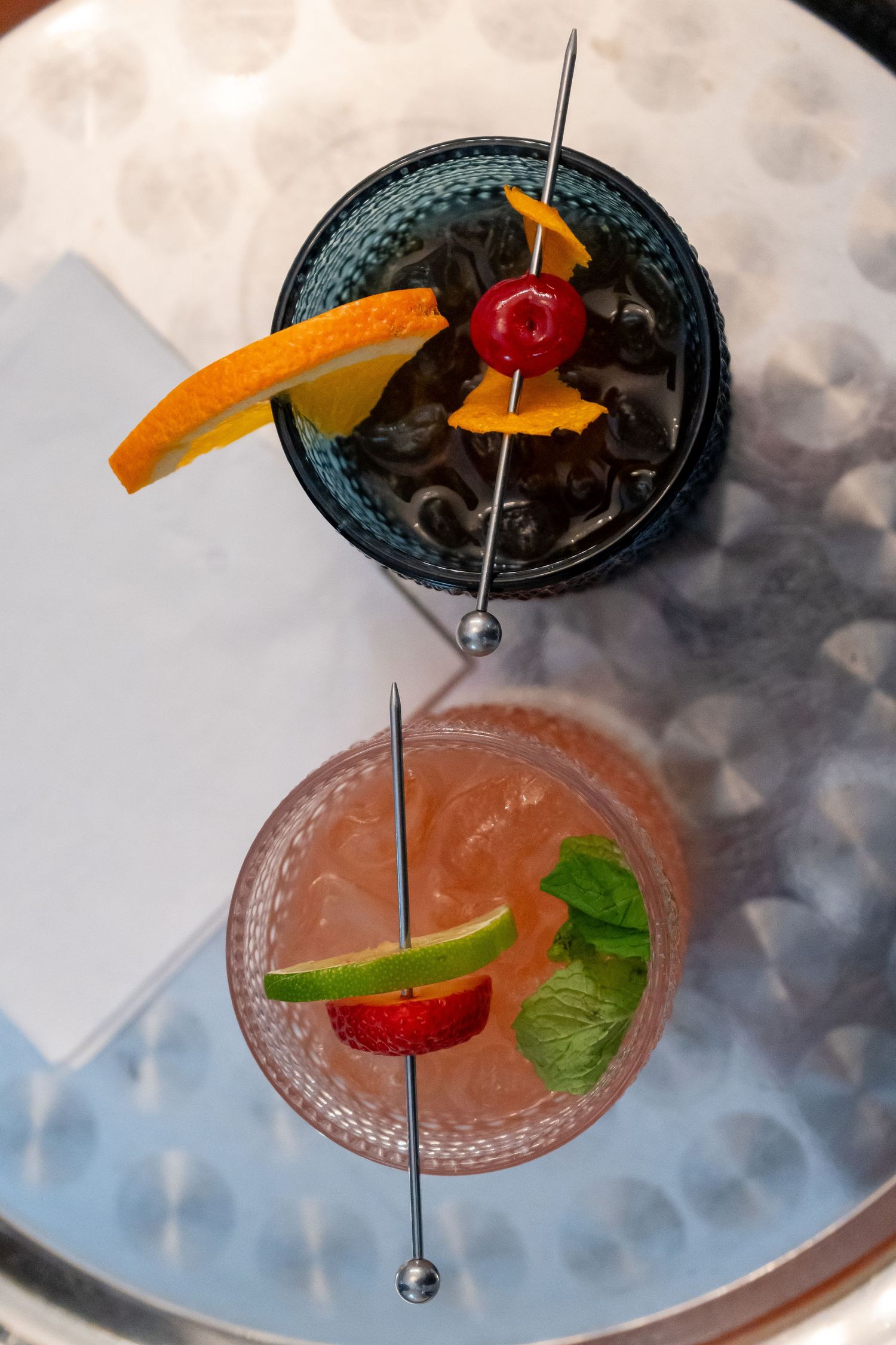 A pink cocktail garnished with orange, cherry and mint leaf on a misty surface.