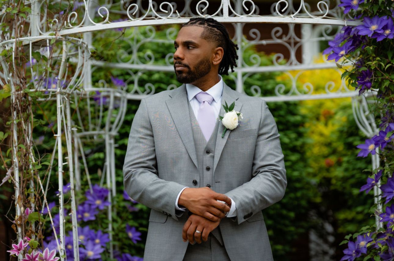 A person in a gray suit stands in a lush garden setting with purple flowers and decorative white archways.