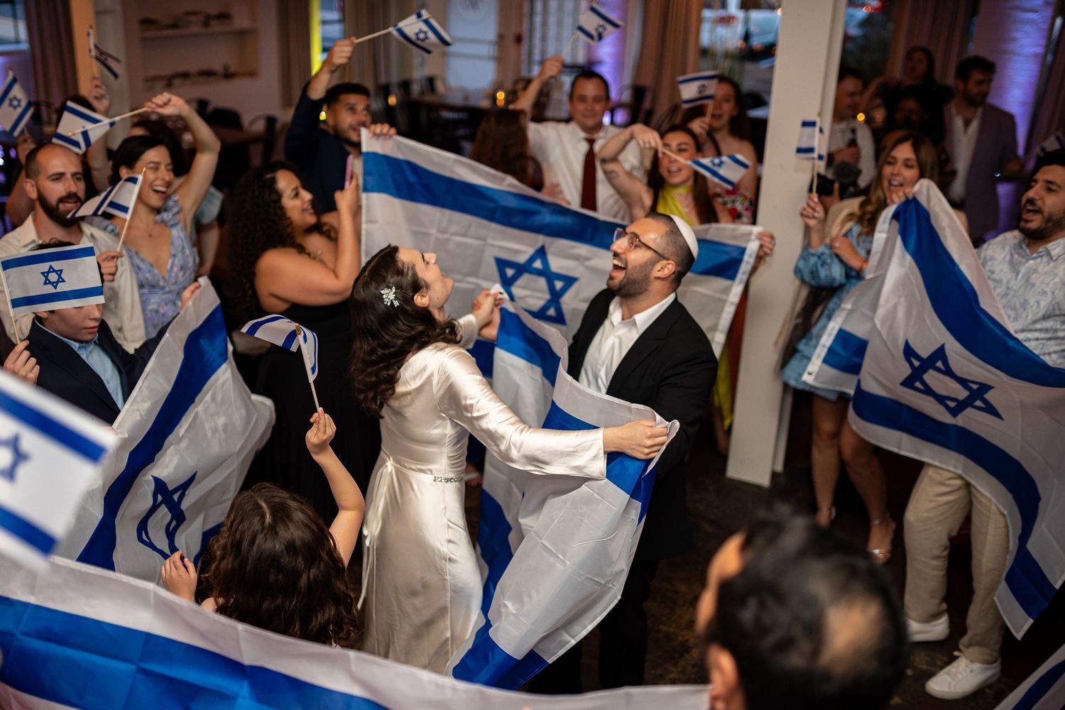 Wedding celebration with guests dancing and waving Israeli flags in a festive atmosphere.