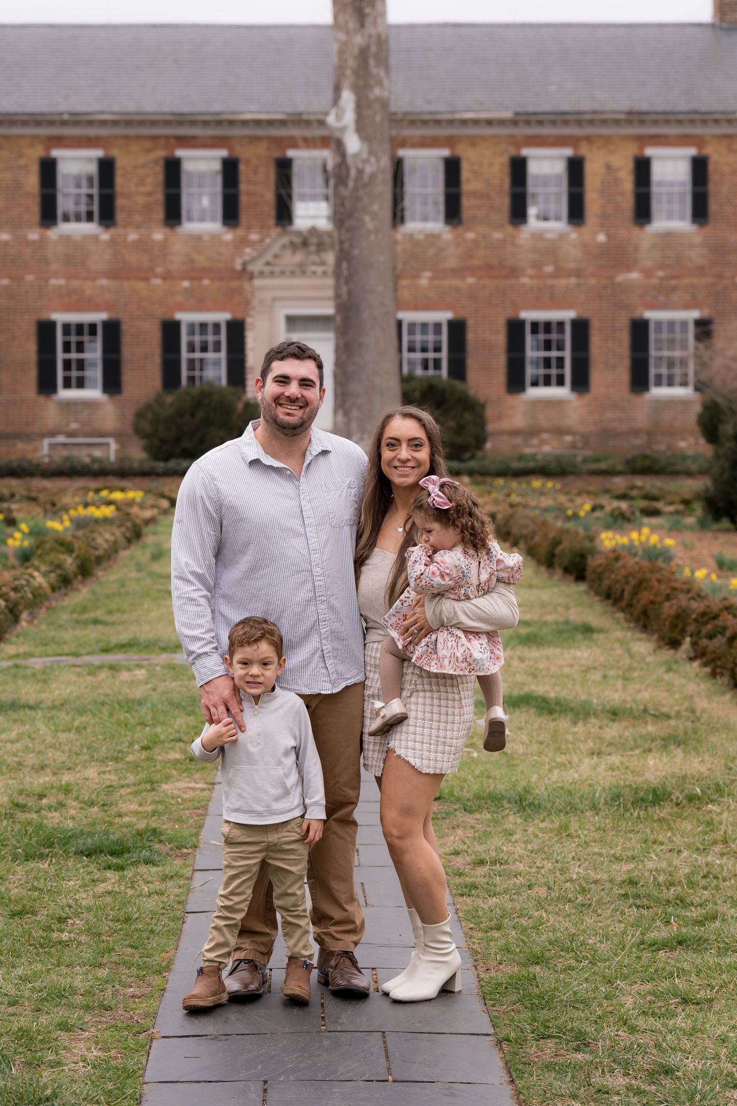 Family poses together on stone steps in front of a brick building wearing coordinated neutral outfits.
