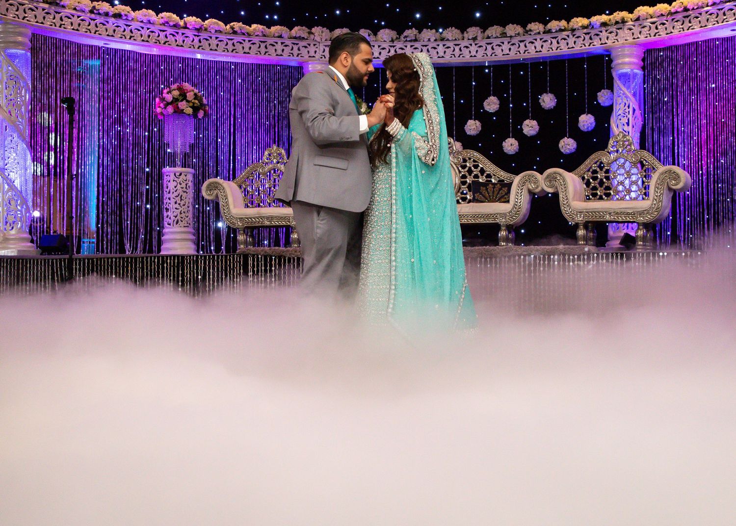 Elegant couple dancing at a luxurious wedding reception with purple decor and ornate seating in the background.