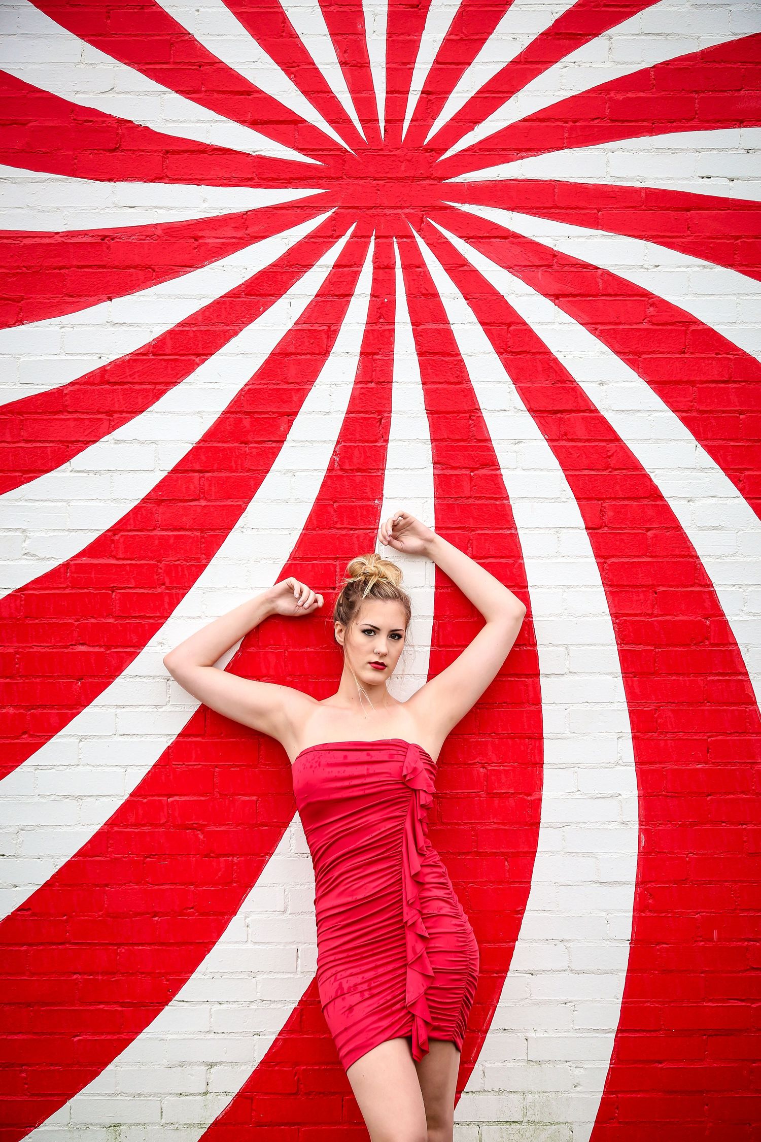 Fashion model poses against striking red and white sunburst pattern wall in vibrant red dress.