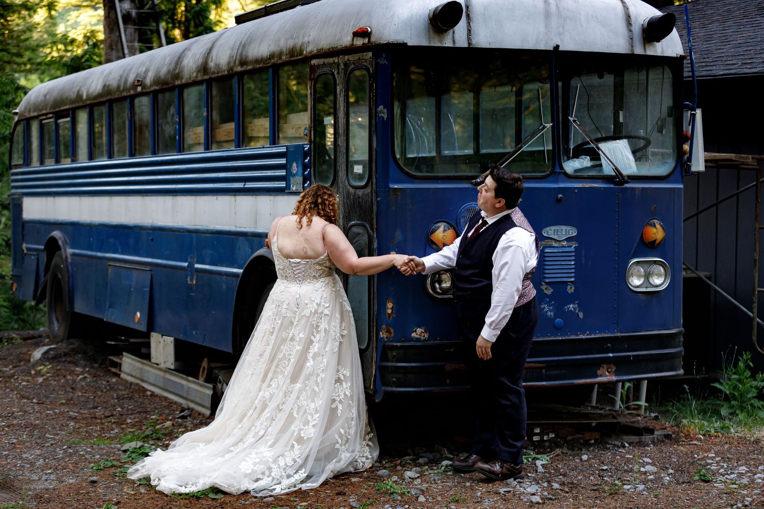 A wedding couple poses creatively around vintage blue buses in a rustic setting.