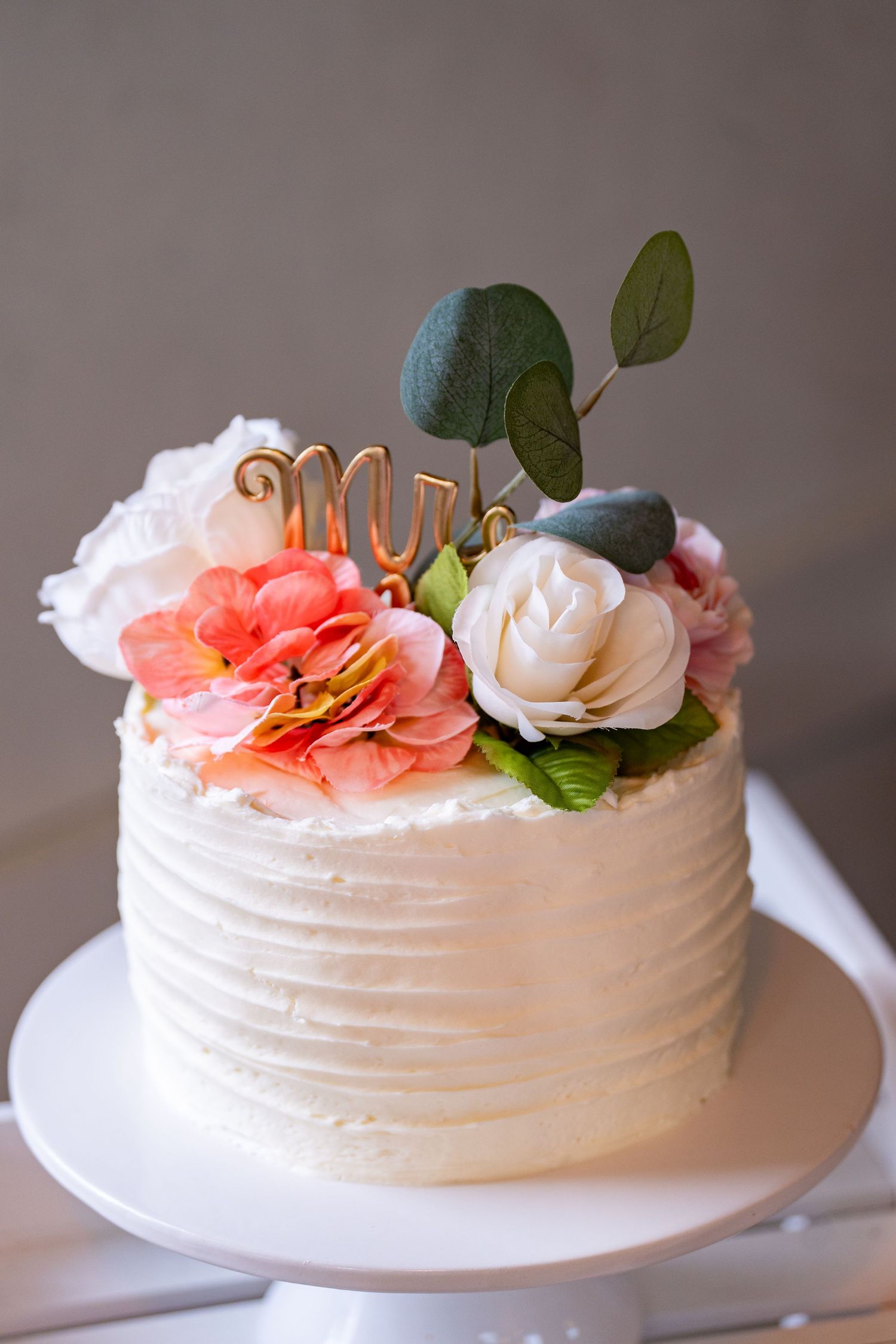 White wedding cake with textured frosting decorated with coral and white flowers and eucalyptus leaves.