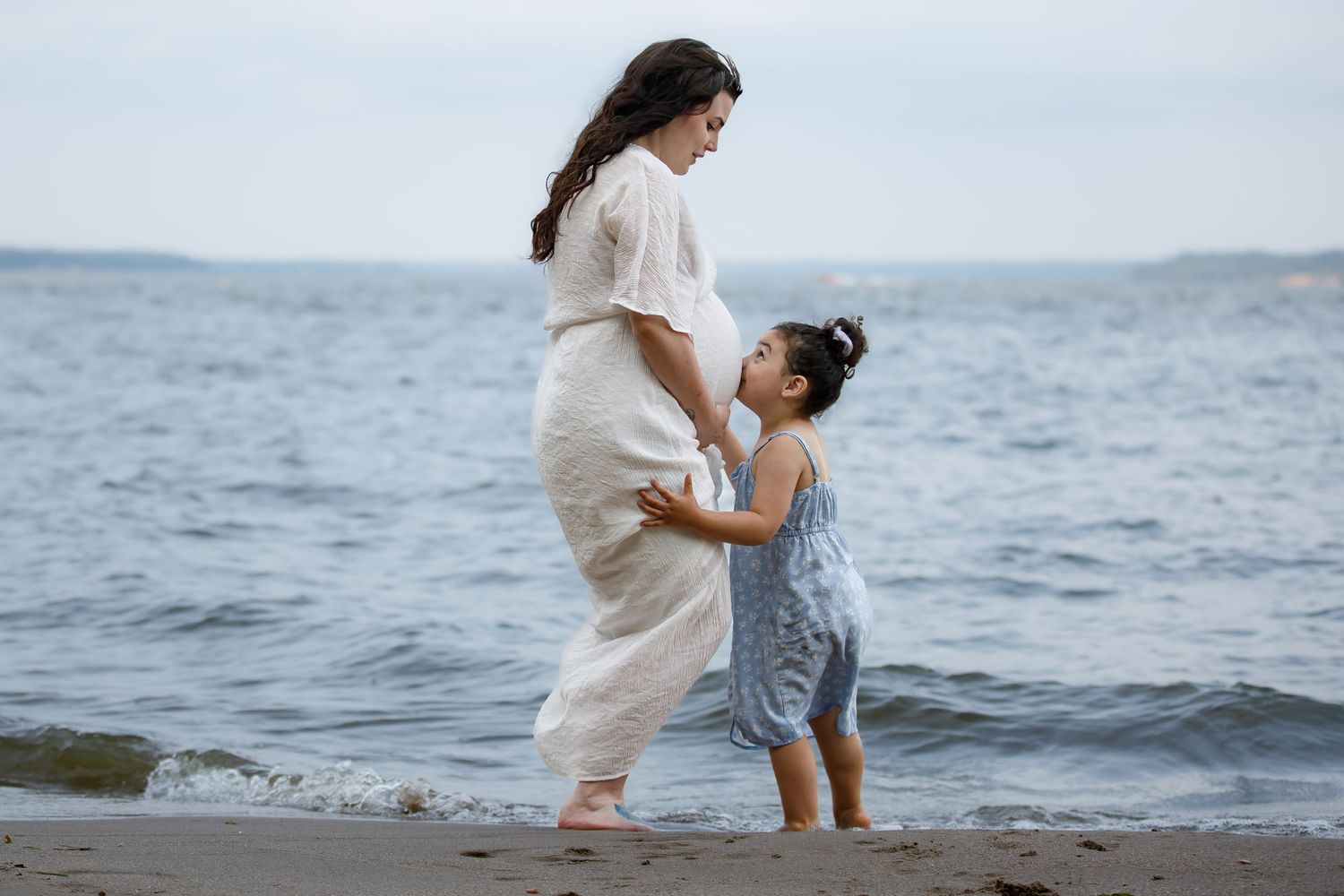 Tender beach moment between parent and child in flowing white and blue dresses.