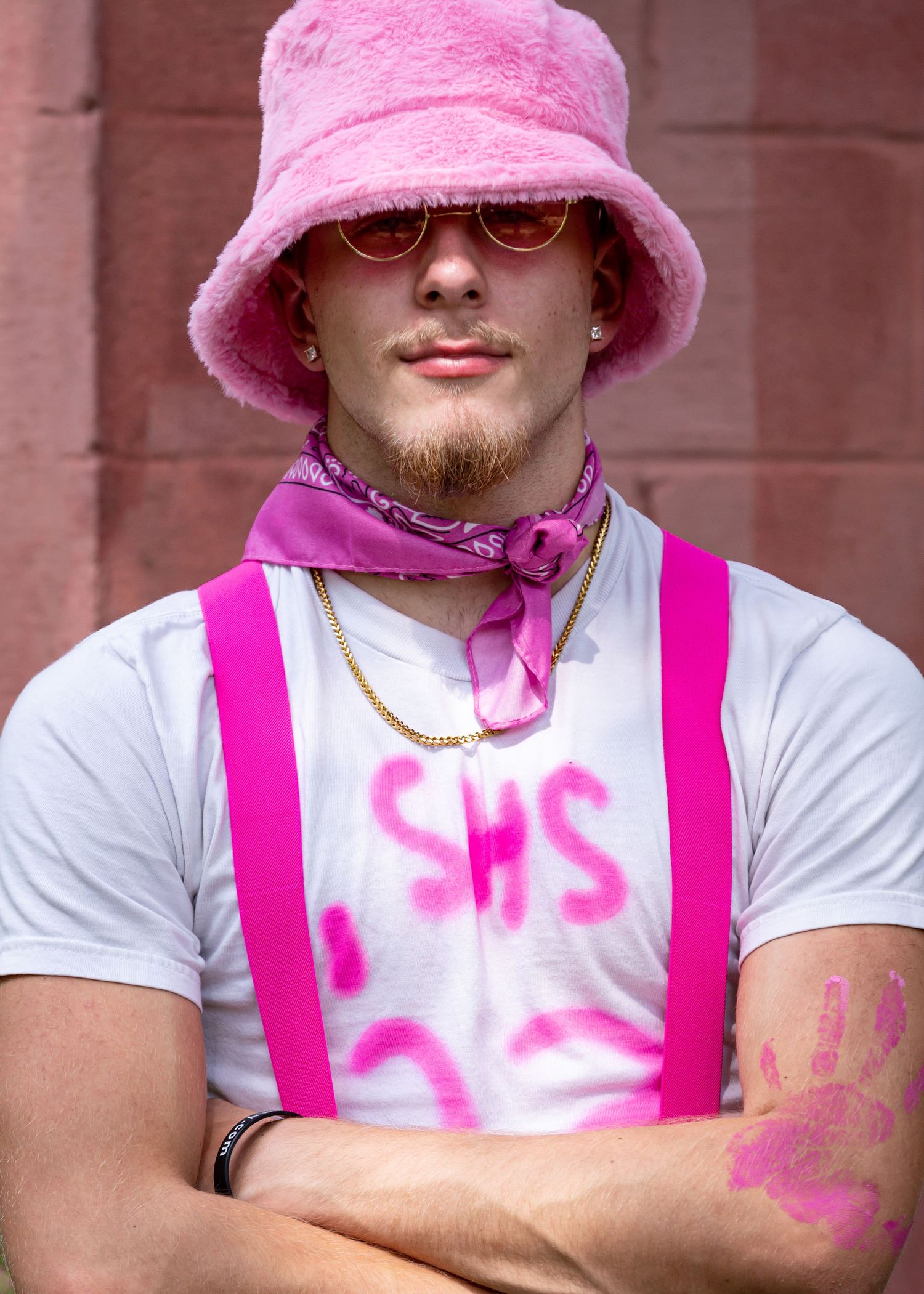 Individual in pink bucket hat and white shirt with neon pink suspenders against brick wall.