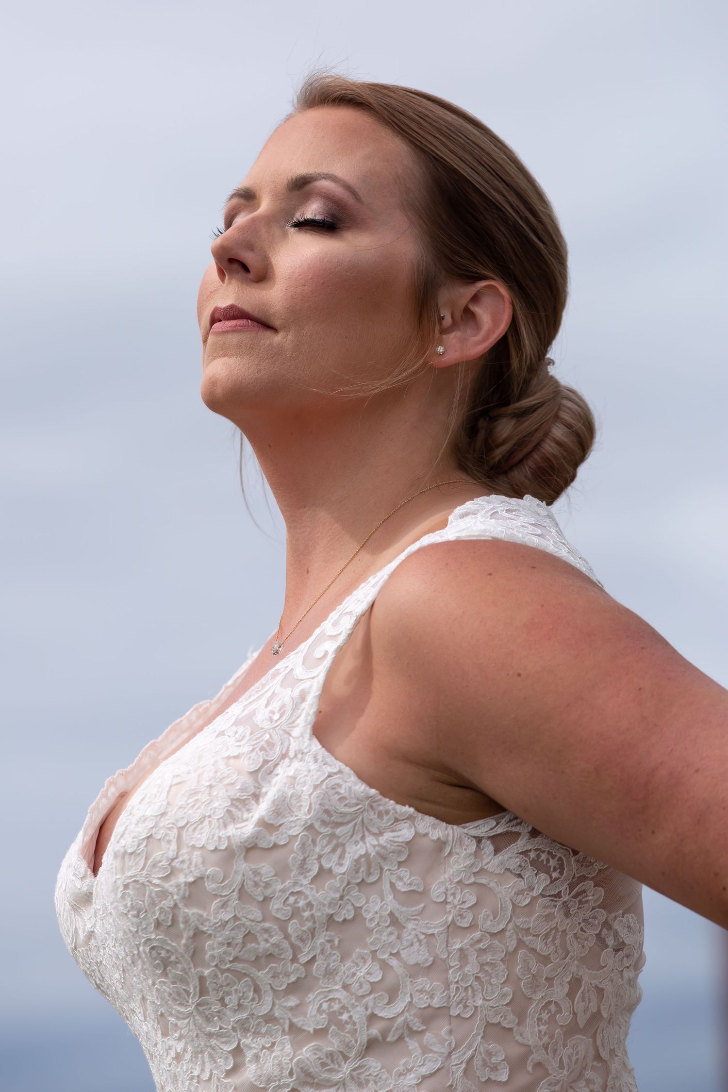 Close up profile portrait of bride in lace dress against cloudy sky.