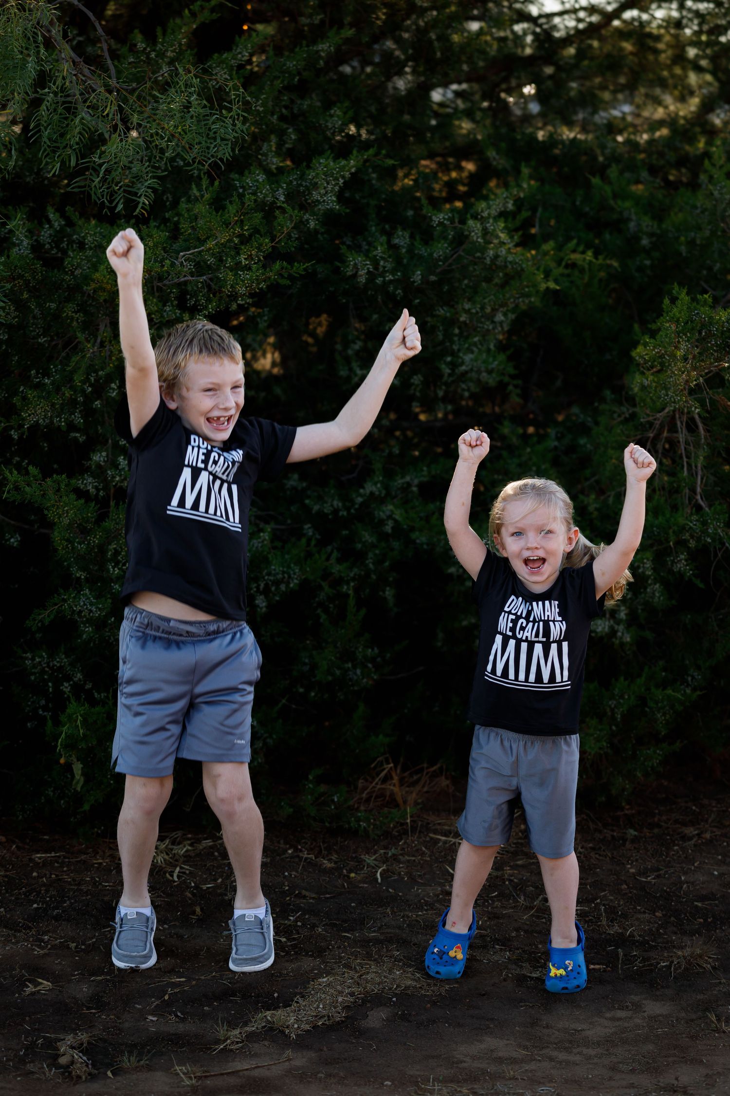 Two children in matching black t-shirts jump with excitement against a dark green background.