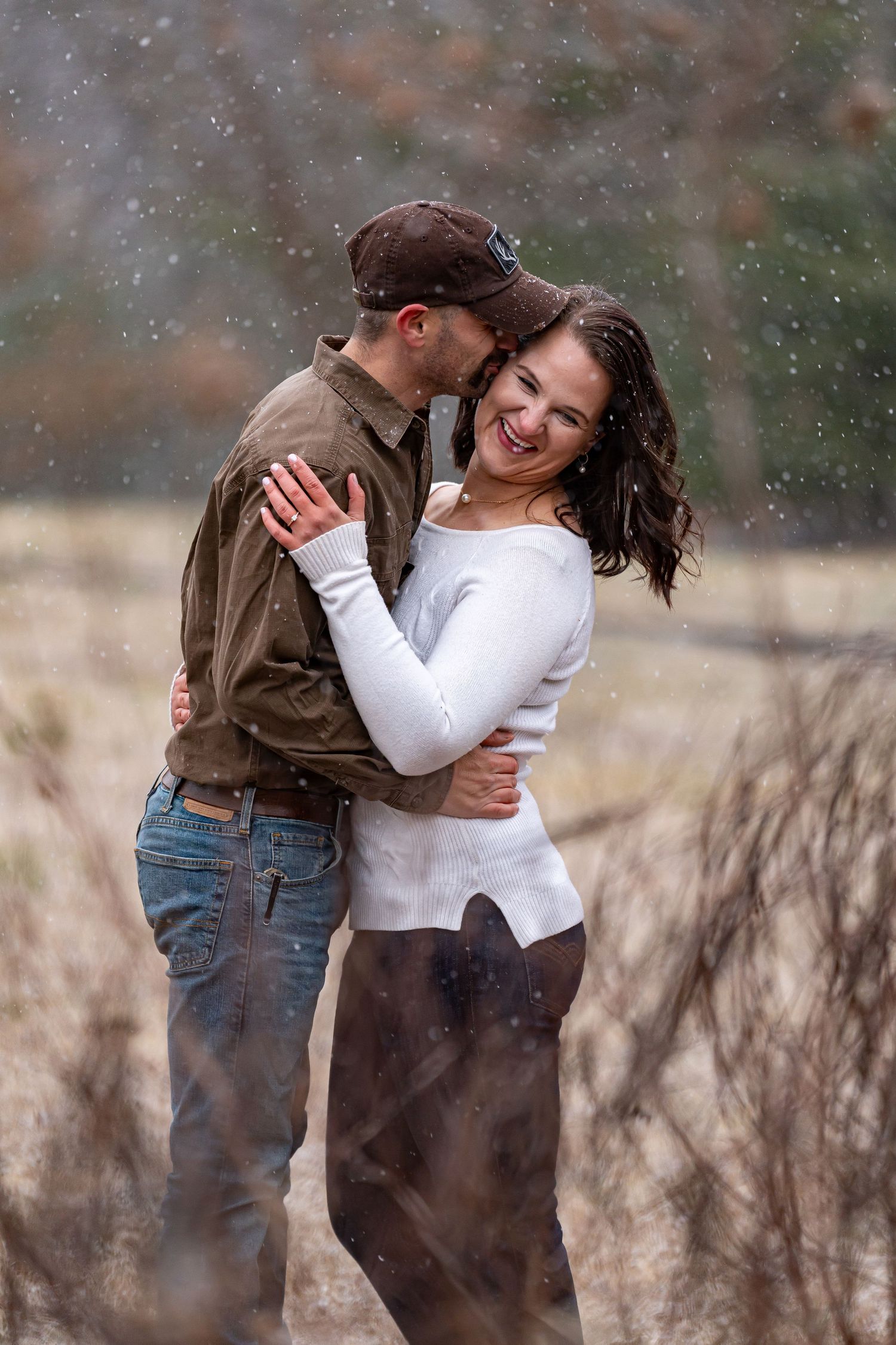 Romantic couple embraces while snow falls in rustic outdoor setting.