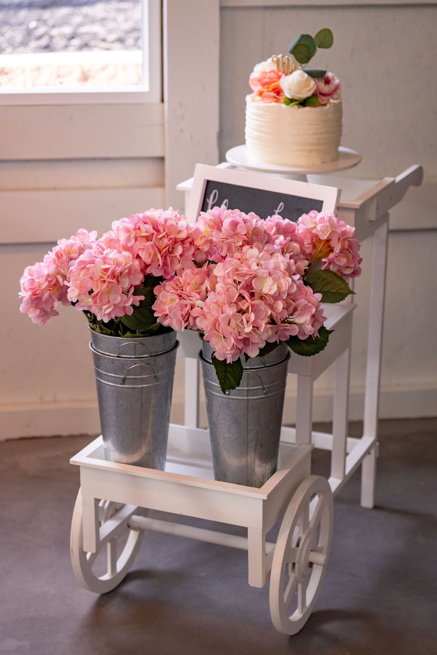 Pink hydrangeas in galvanized metal buckets displayed on white decorative cart with small cake.