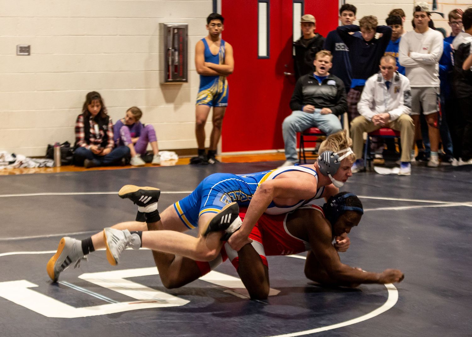 Wrestling match in progress with competitors in red and blue uniforms on indoor mat.