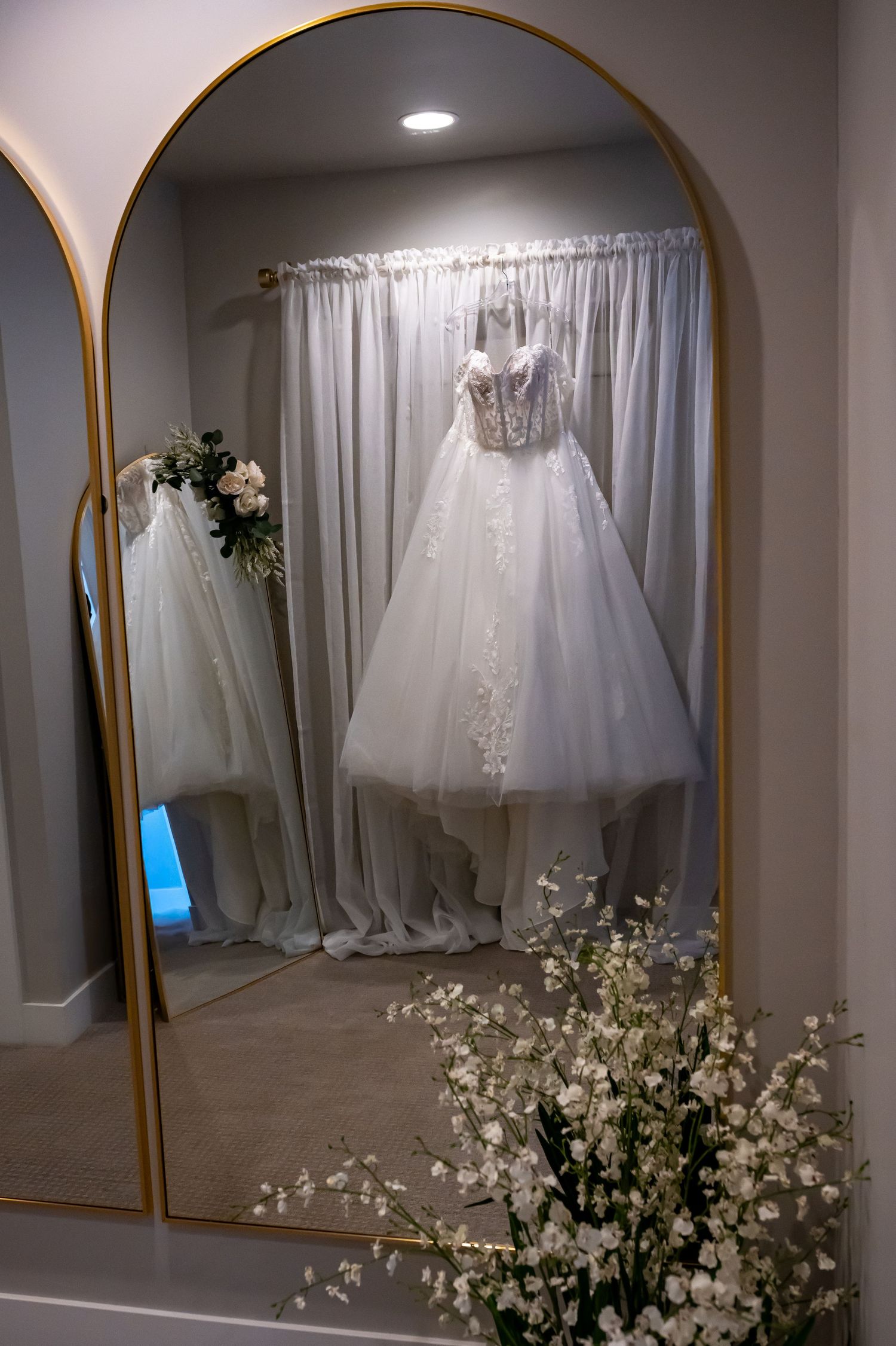 Multiple wedding dresses hanging on display with white curtains and floral decorations.