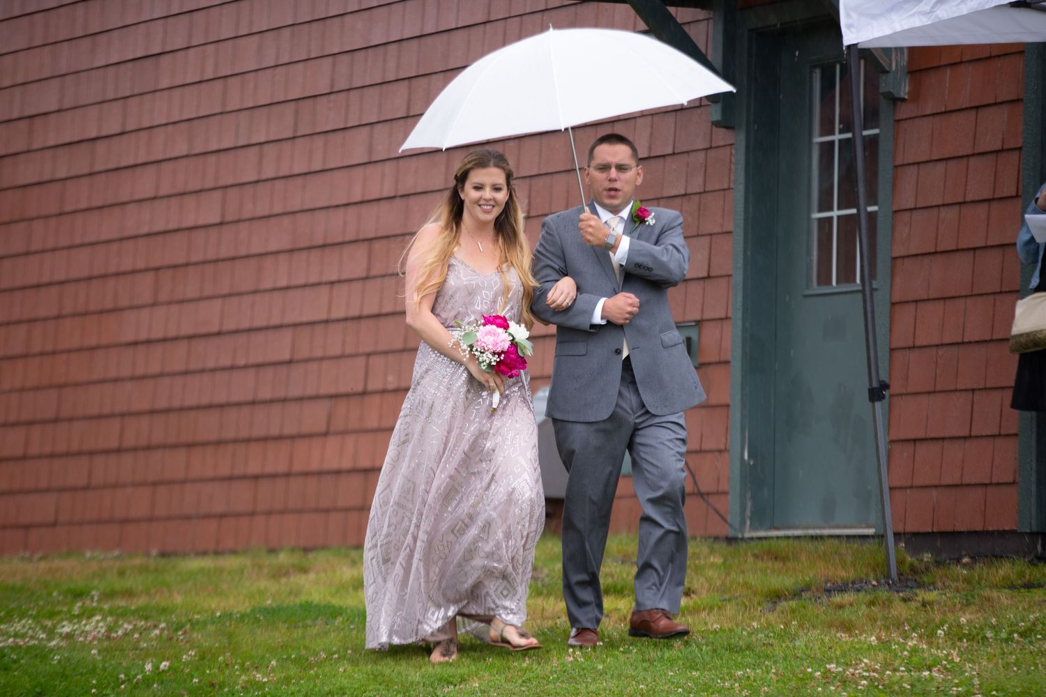 A couple walking together under a white umbrella outside a rustic cedar-sided building on their wedding day.