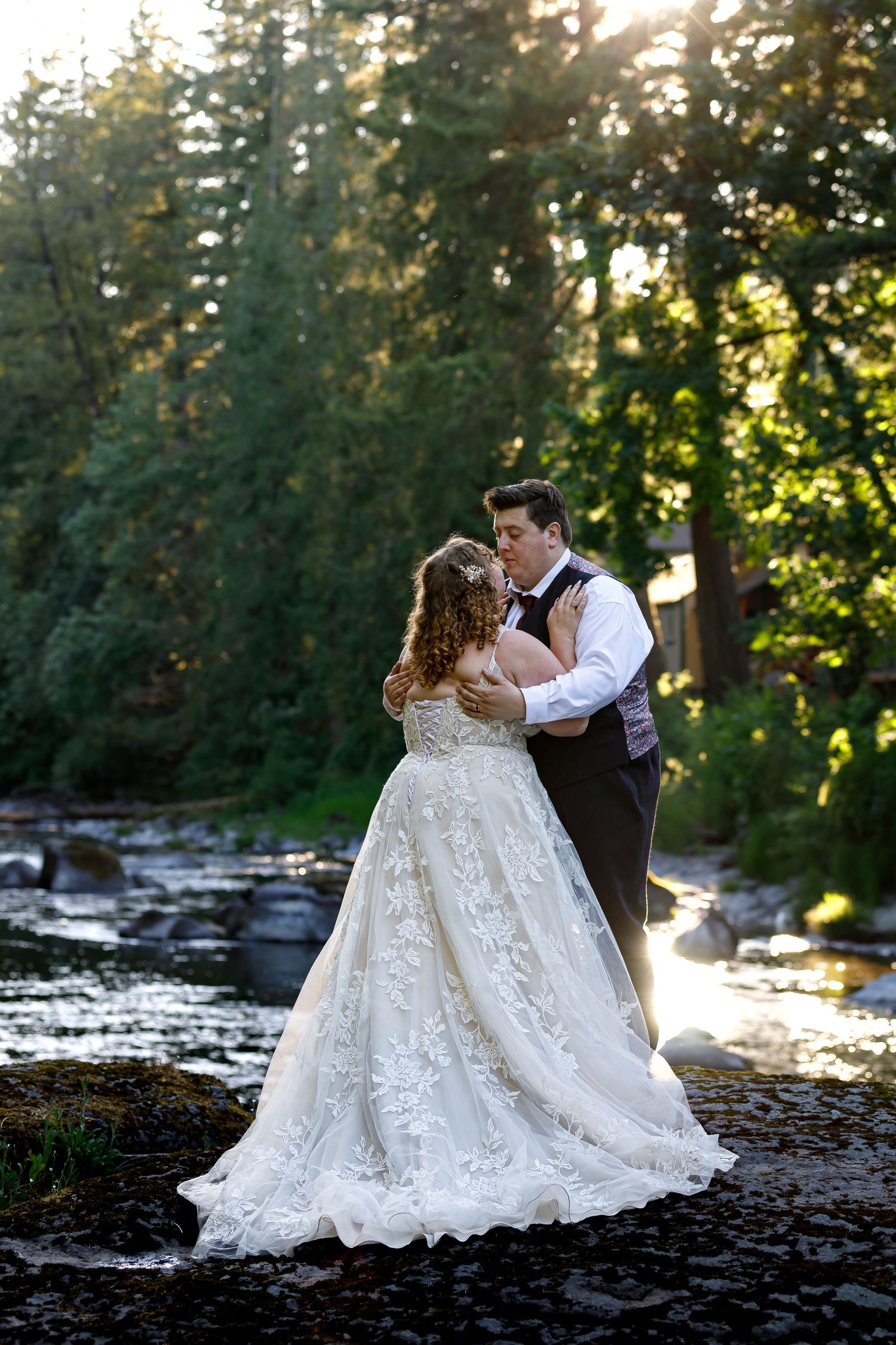 Bride and groom dancing by forest stream during golden hour.