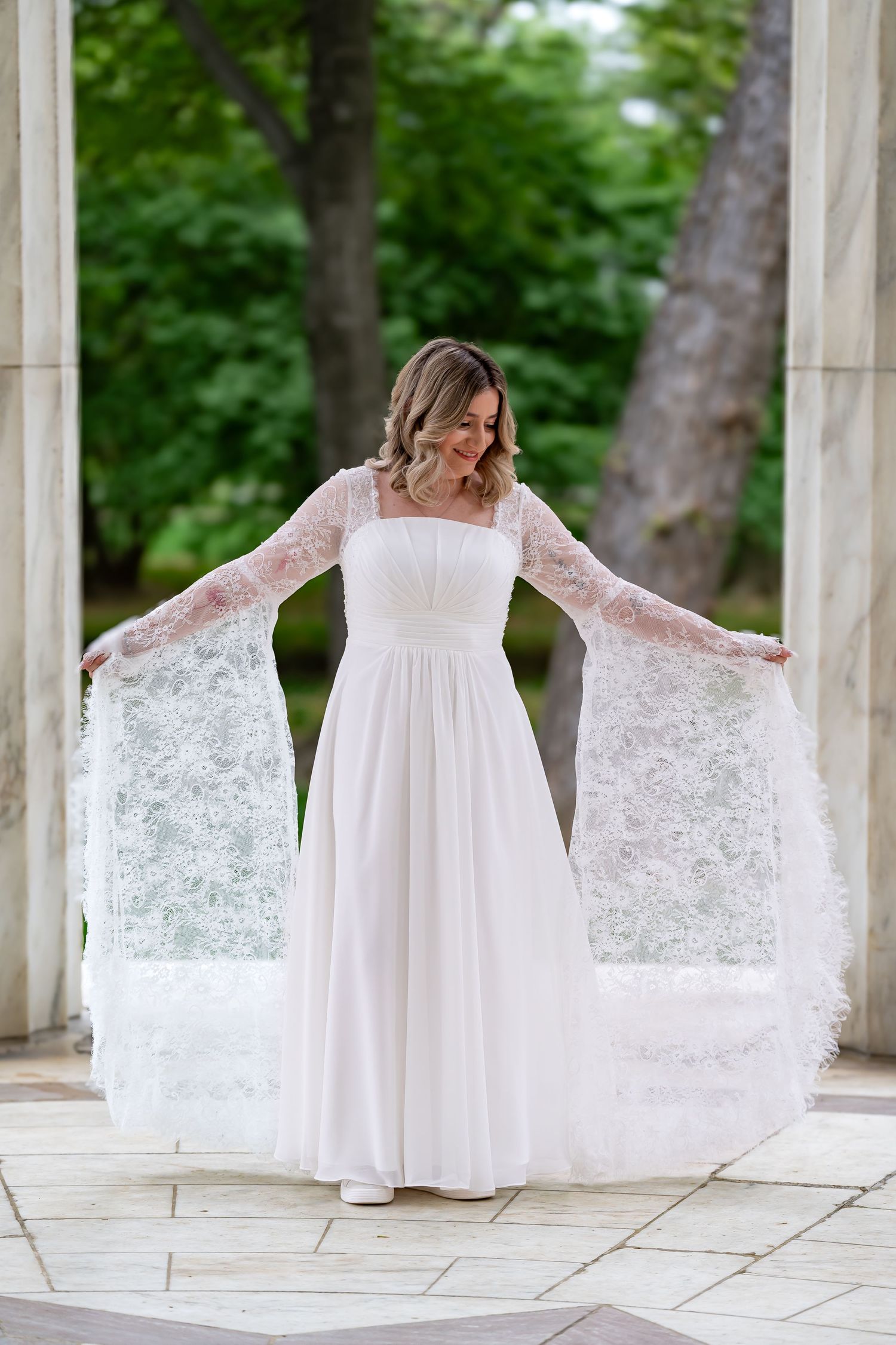 Bride in flowing white lace dress poses elegantly between marble columns outdoors.