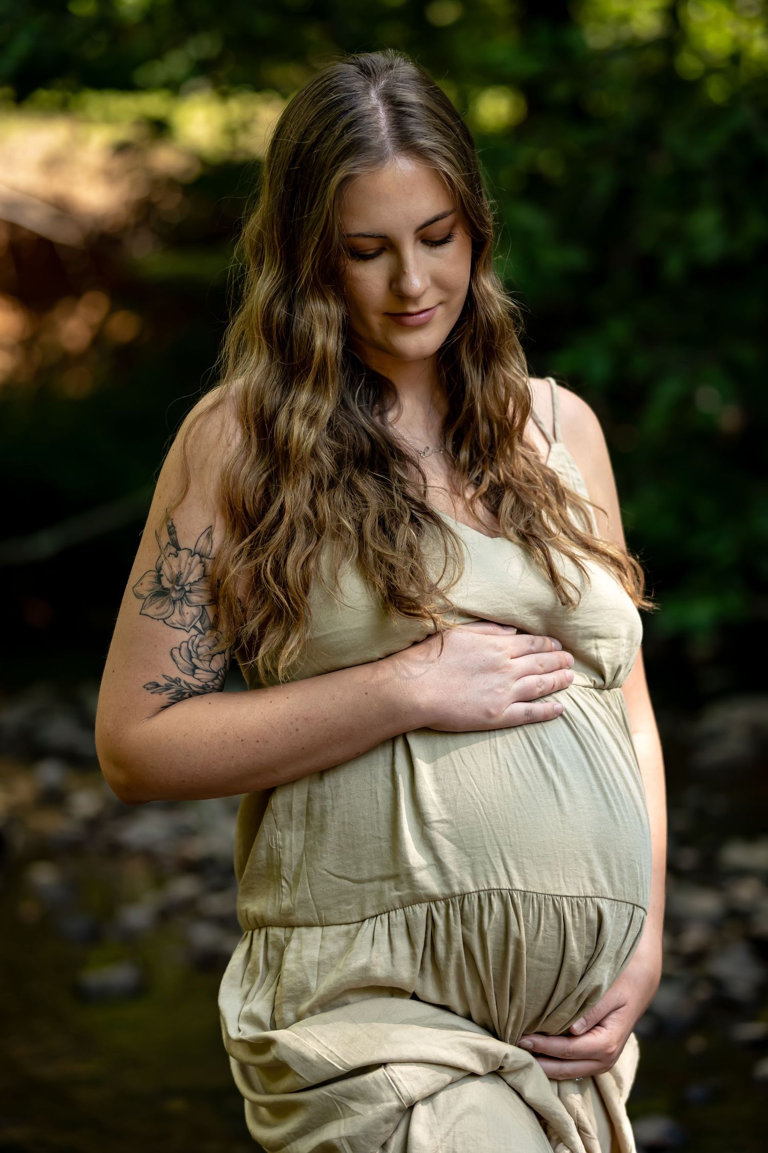 Expectant mother in flowing beige dress poses tenderly in outdoor setting.