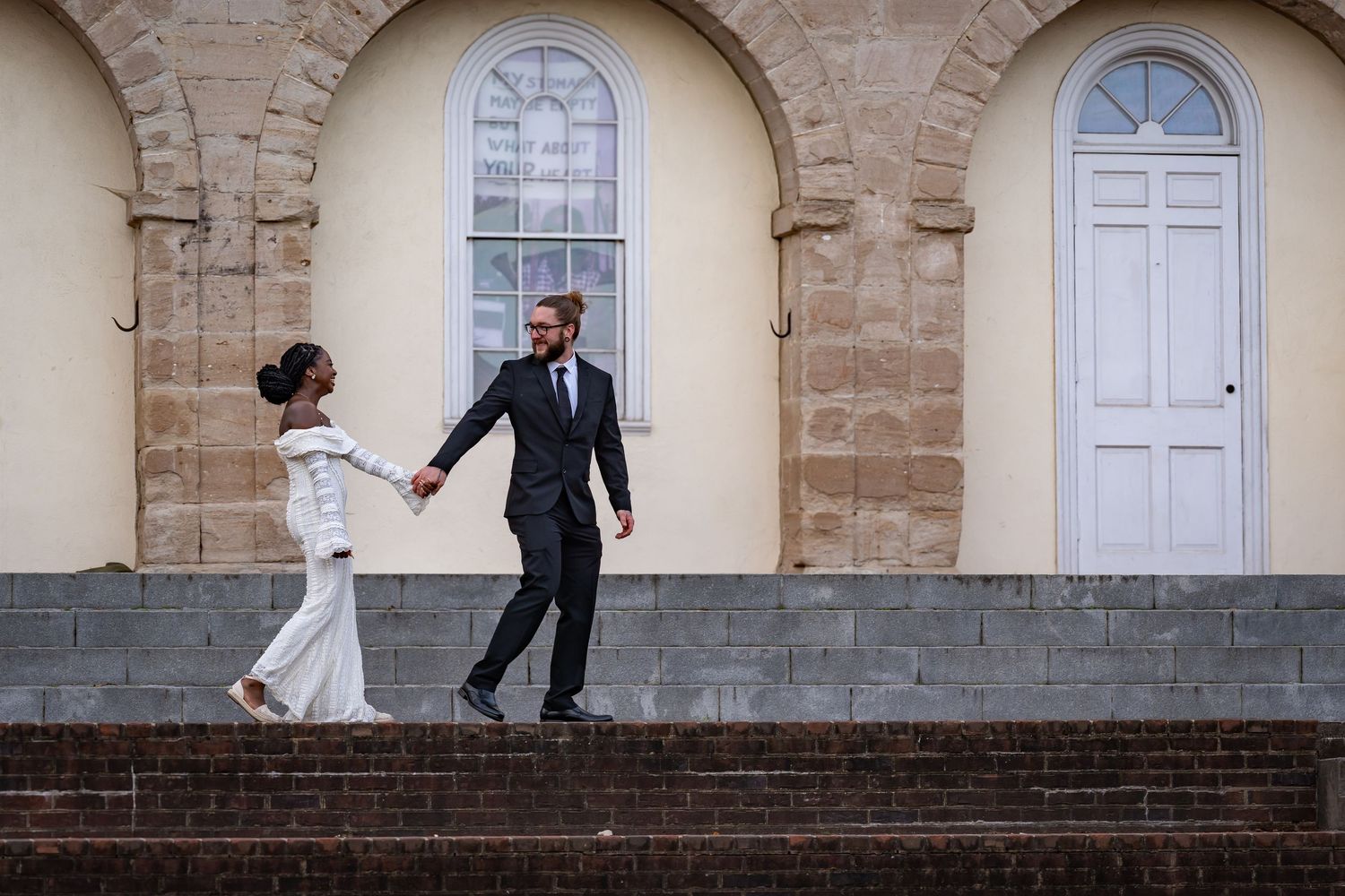 A couple in wedding attire dances playfully on stone steps in front of an arched doorway.